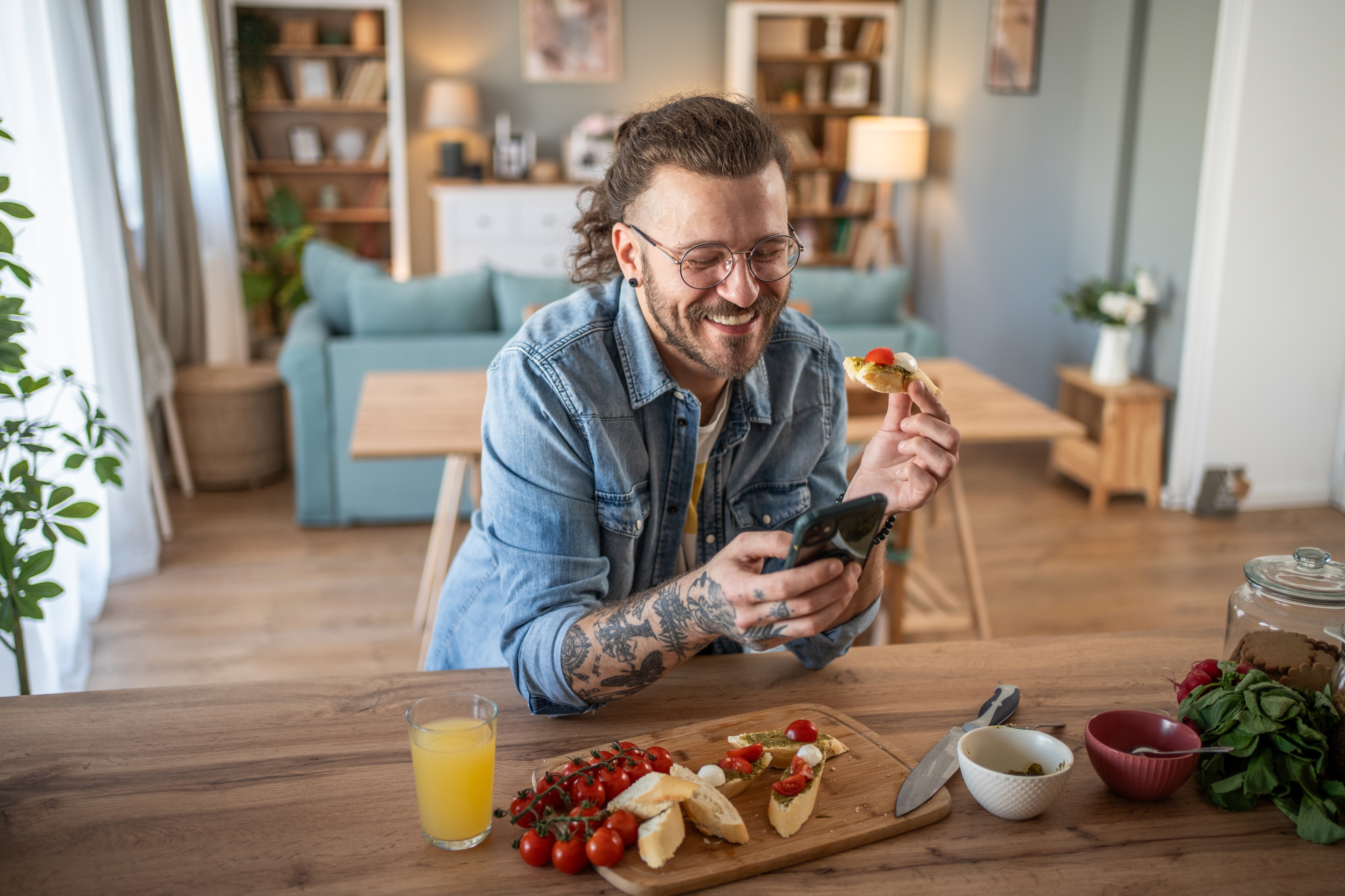 Man with beard and tattoos, wearing glasses and a denim jacket, smiles while looking at his phone and eating bruschetta at a kitchen table