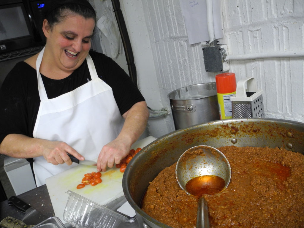 Person in kitchen slicing vegetables near a large pot of chili, smiling while preparing food