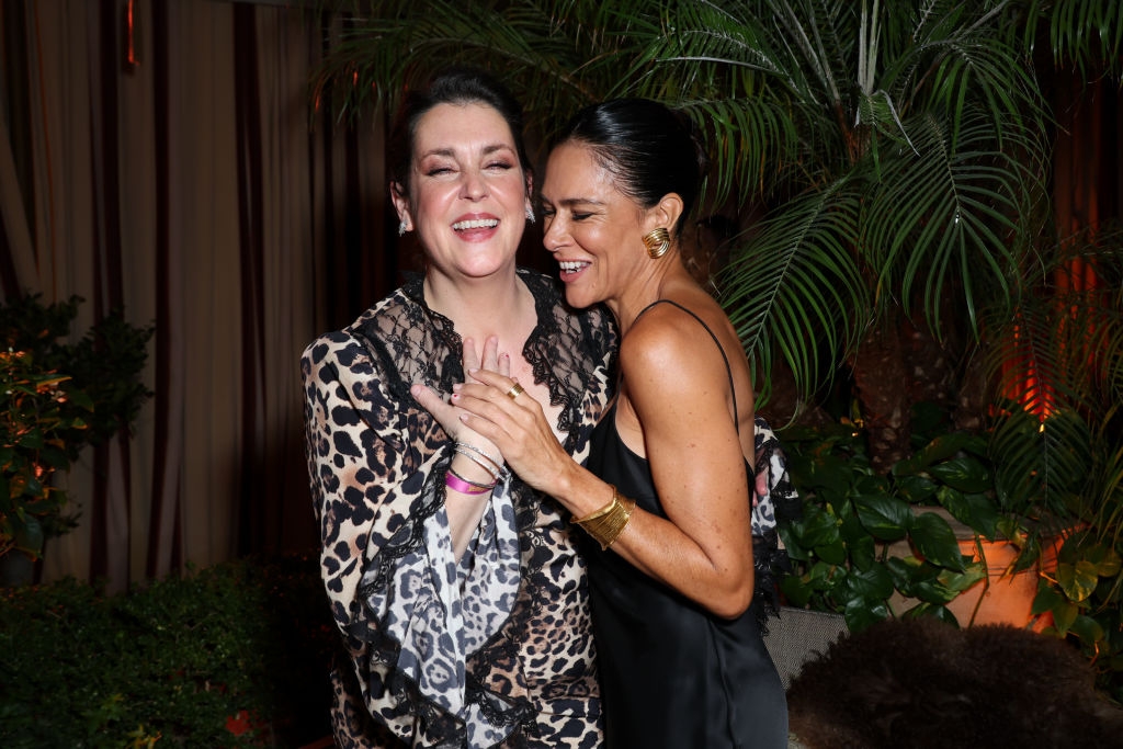Two women smiling, one in a leopard print dress, the other in a black slip dress, in a lively, plant-filled setting
