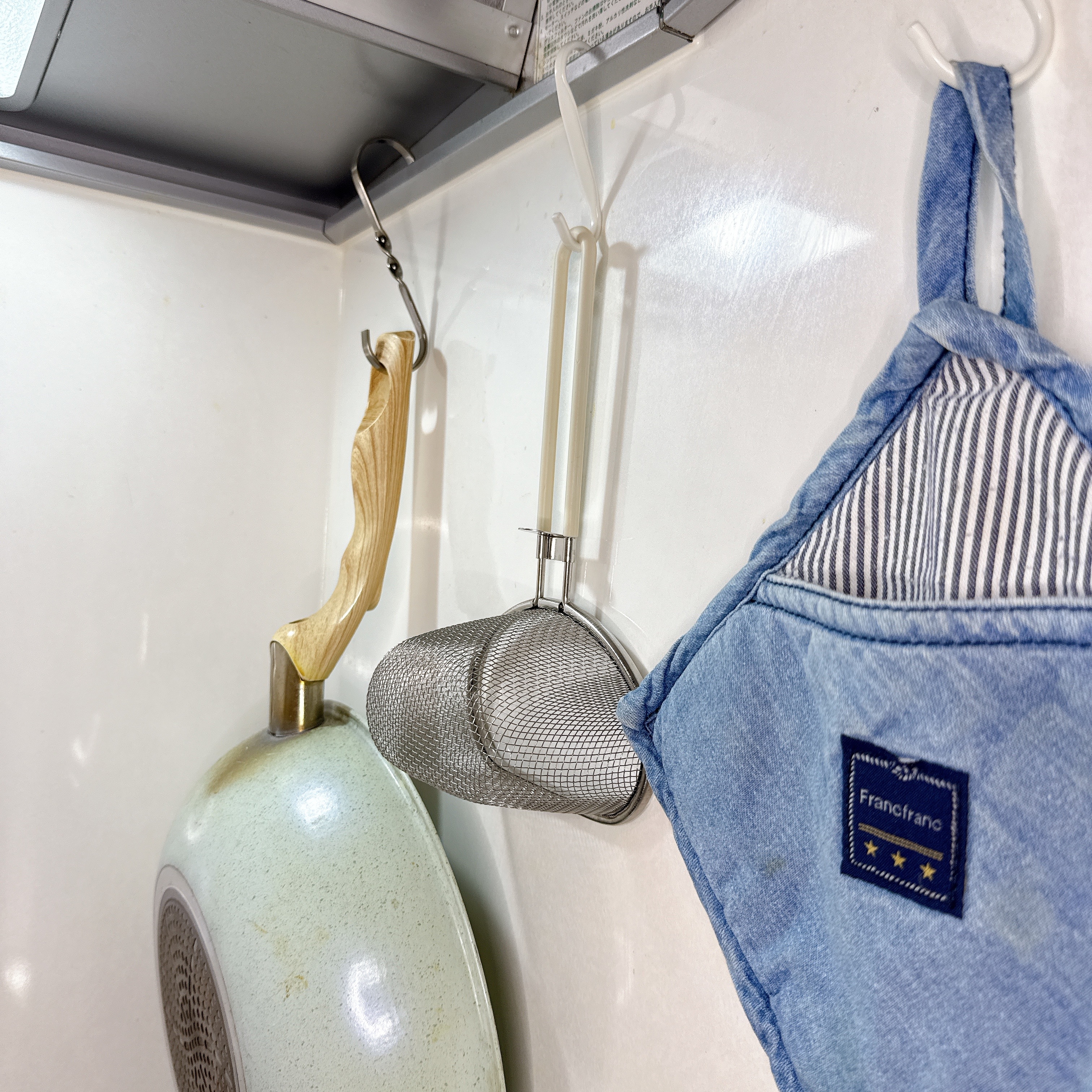 キッチンの壁にフライパン、茶こし、青い鍋つかみが掛けられた収納アイデア。