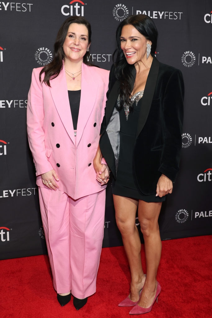 Two people pose on a red carpet; one in a pink suit, the other in a black dress with a jacket