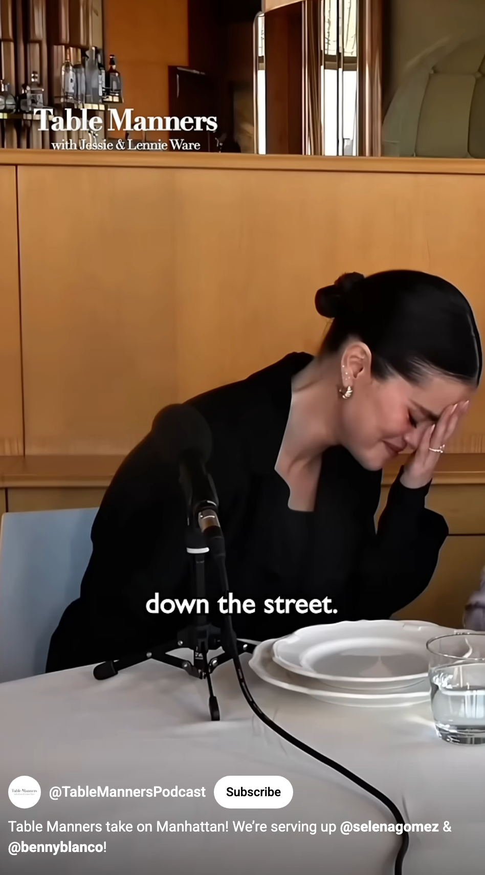 Person at a podcast table smiling with head in hand, wearing a sleek outfit, and a plate in front of them. Text: &quot;down the street.&quot;