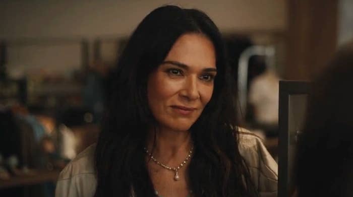 Person with long dark hair smiling warmly, wearing a light top and a pearl necklace, in an indoor setting