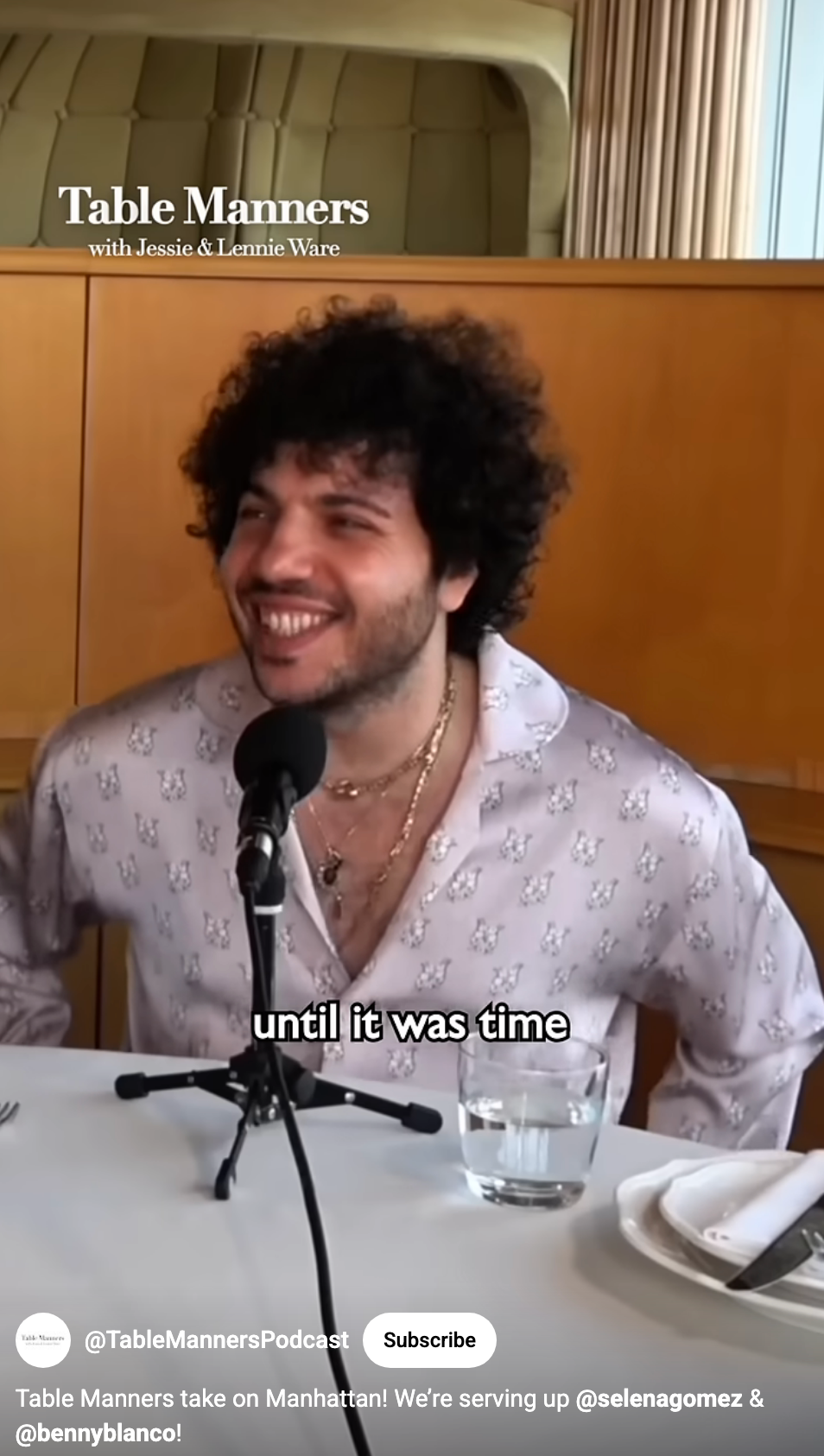 A smiling person sits at a table with microphones for a podcast interview, wearing a patterned shirt