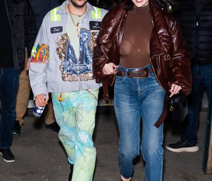 Two people walking happily. The woman wears a brown leather jacket and jeans, the man wears a graphic jacket and tie-dye pants