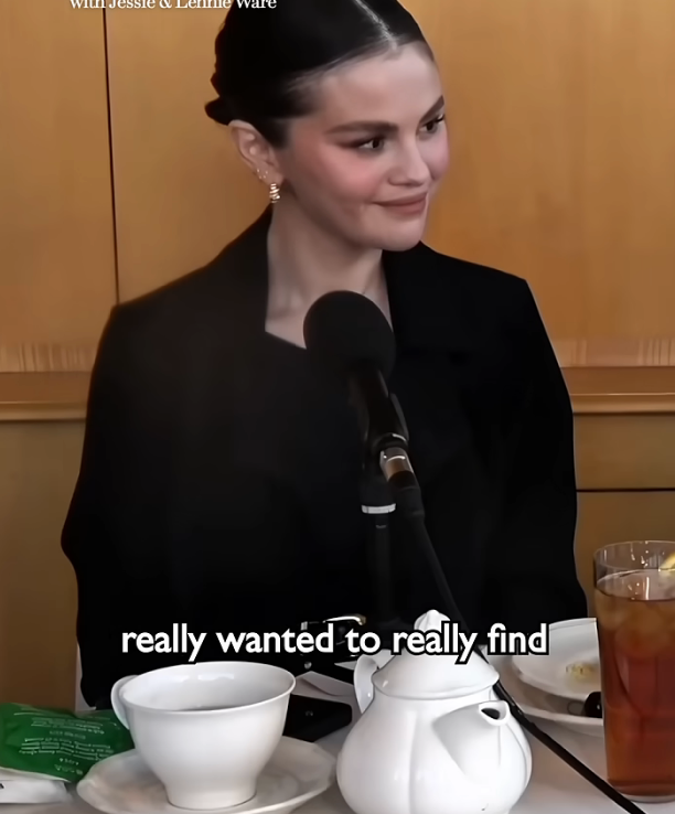 Person in a black jacket speaking at a podcast, seated at a table with tea, facing a microphone