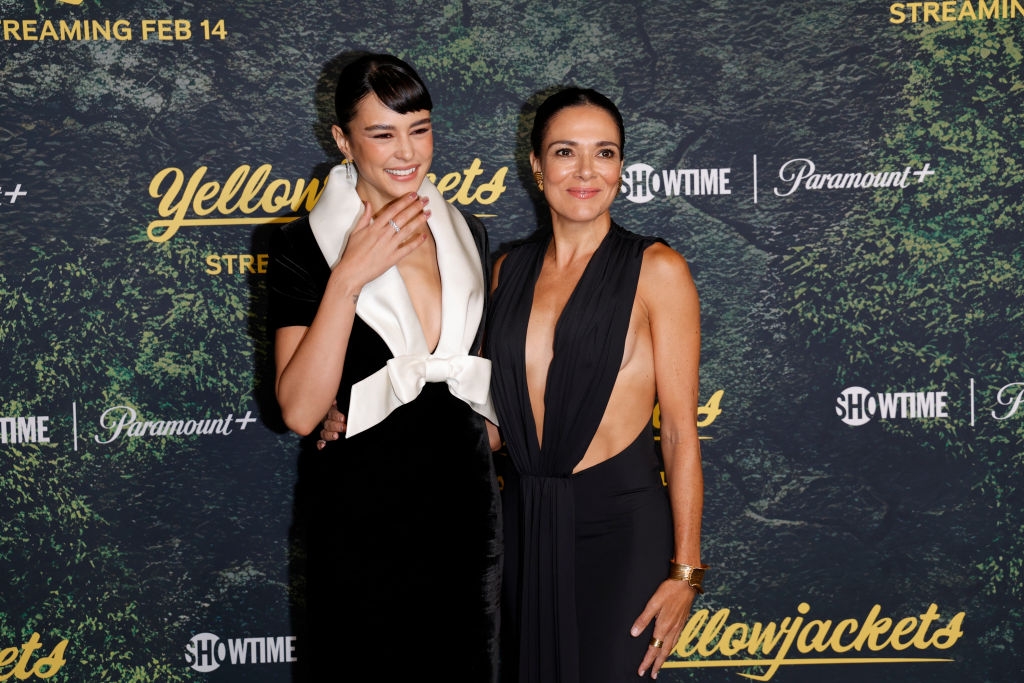 Two women stand on a Showtime event's red carpet. One wears a sleeveless dress with a white bow; the other wears a deep v-neck dress