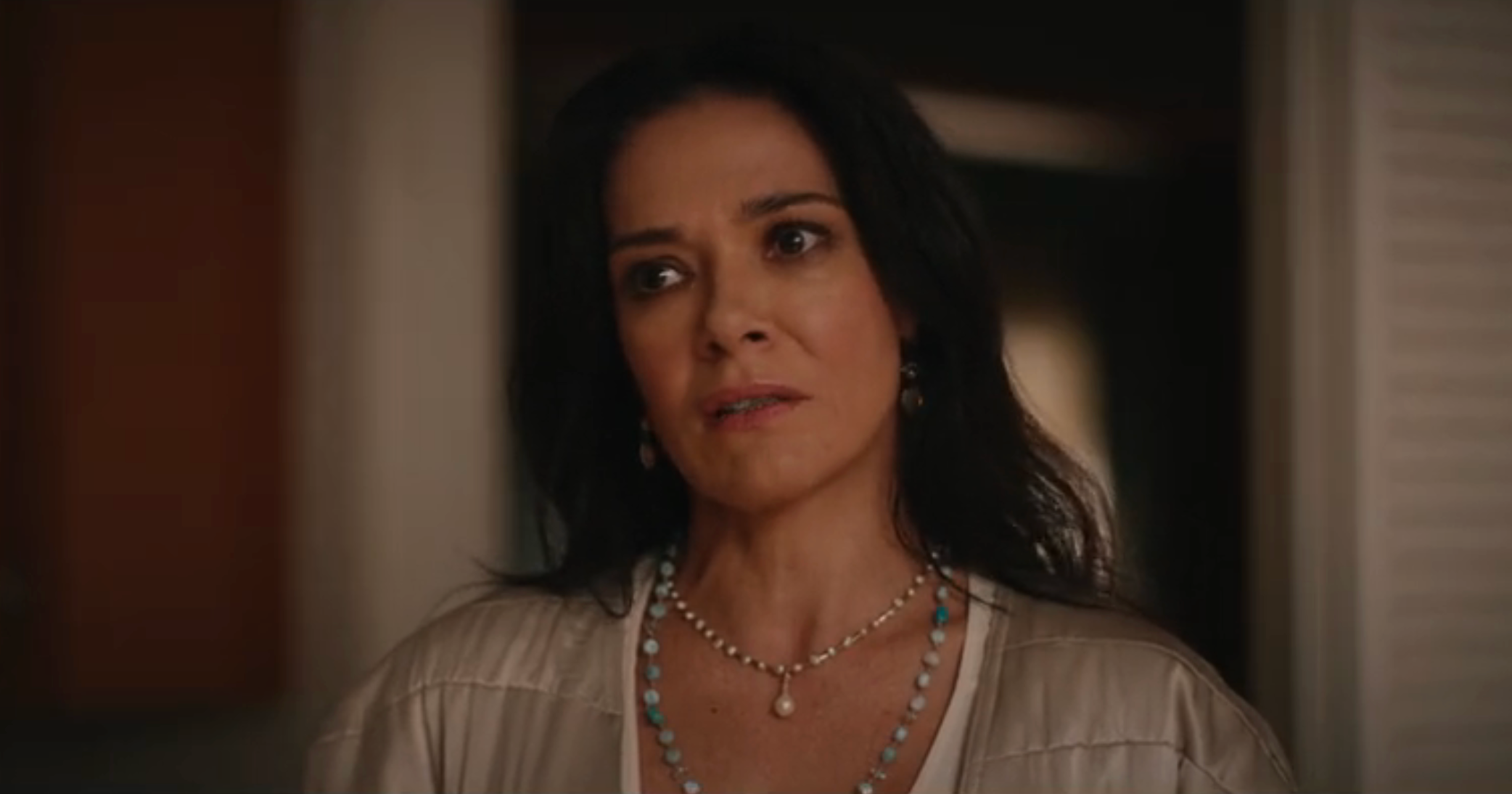A person looks thoughtful, wearing a pearl necklace and earrings, dressed in a casual top