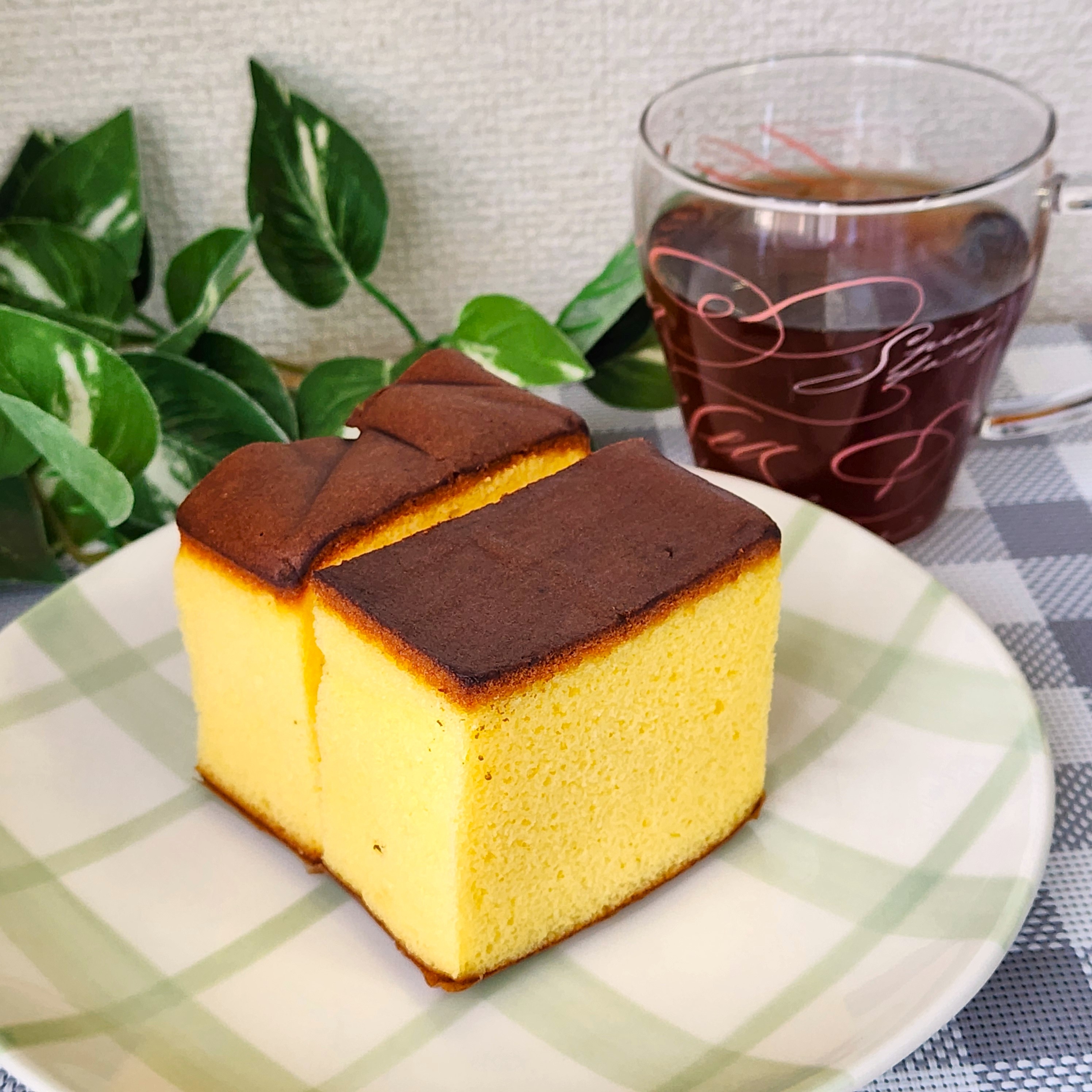 プレートにカステラの切り分けが2つと、隣にレトロなガラスカップに入った紅茶が置かれている。背景に植物。