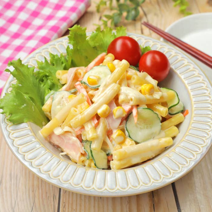マカロニサラダが白い皿に盛られており、キュウリ、コーン、トマト、レタスが添えられています。