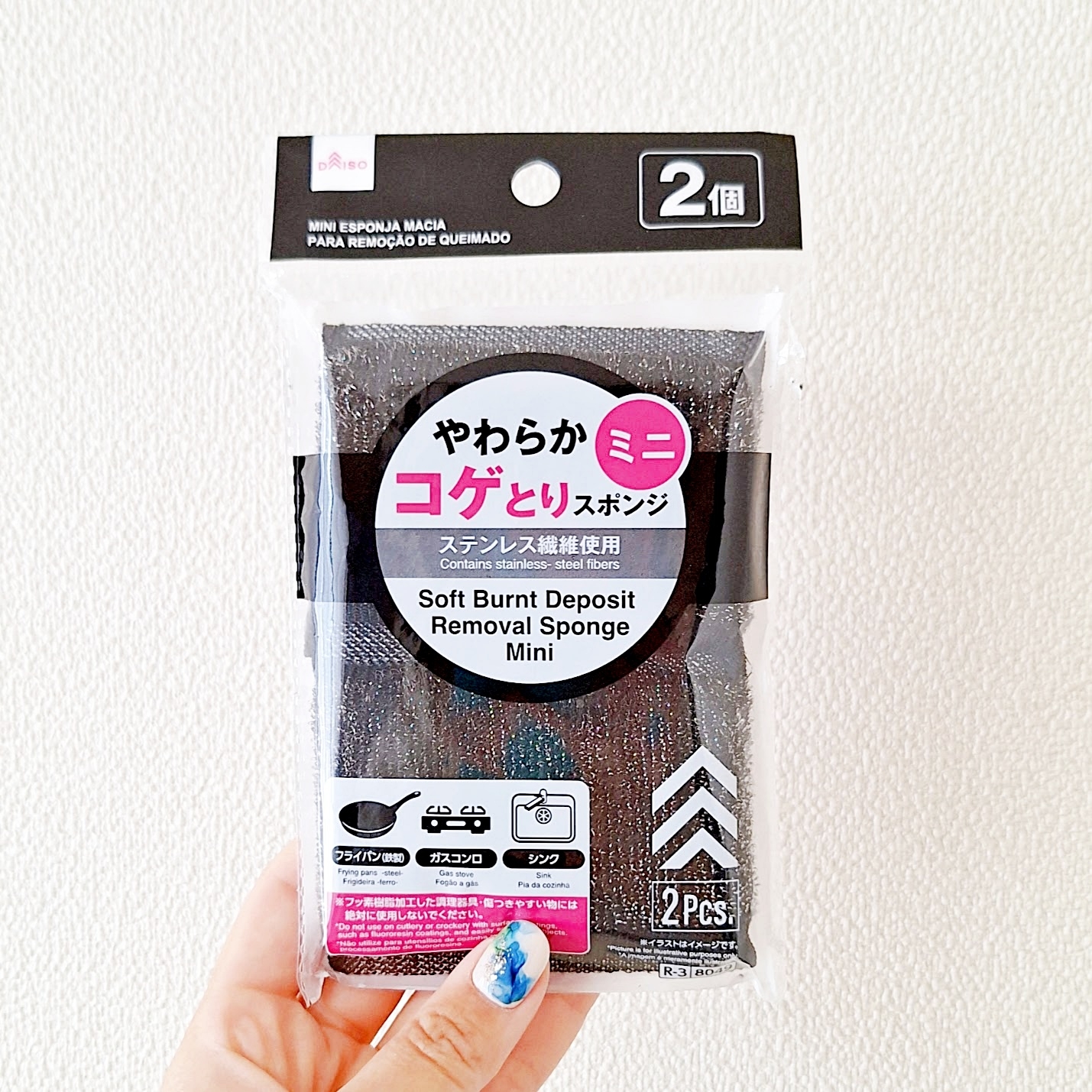 商品パッケージ:焦げ取りスポンジ、2個入り、ステンレス製品用。手で持たれている。