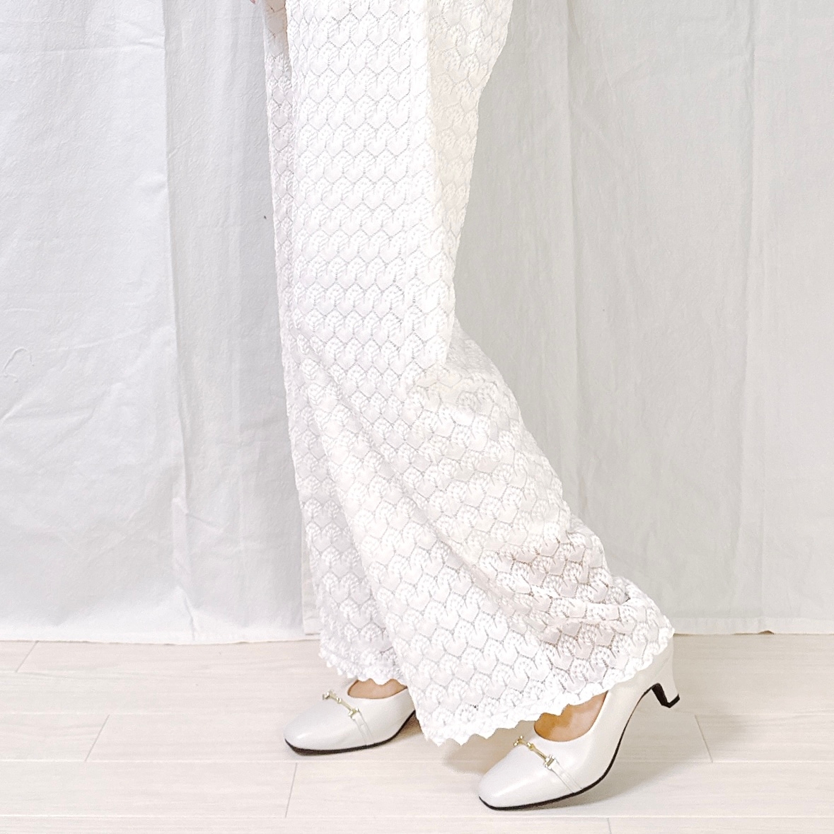 Person wearing textured, floor-length lace pants and white heeled shoes stands in front of a plain backdrop