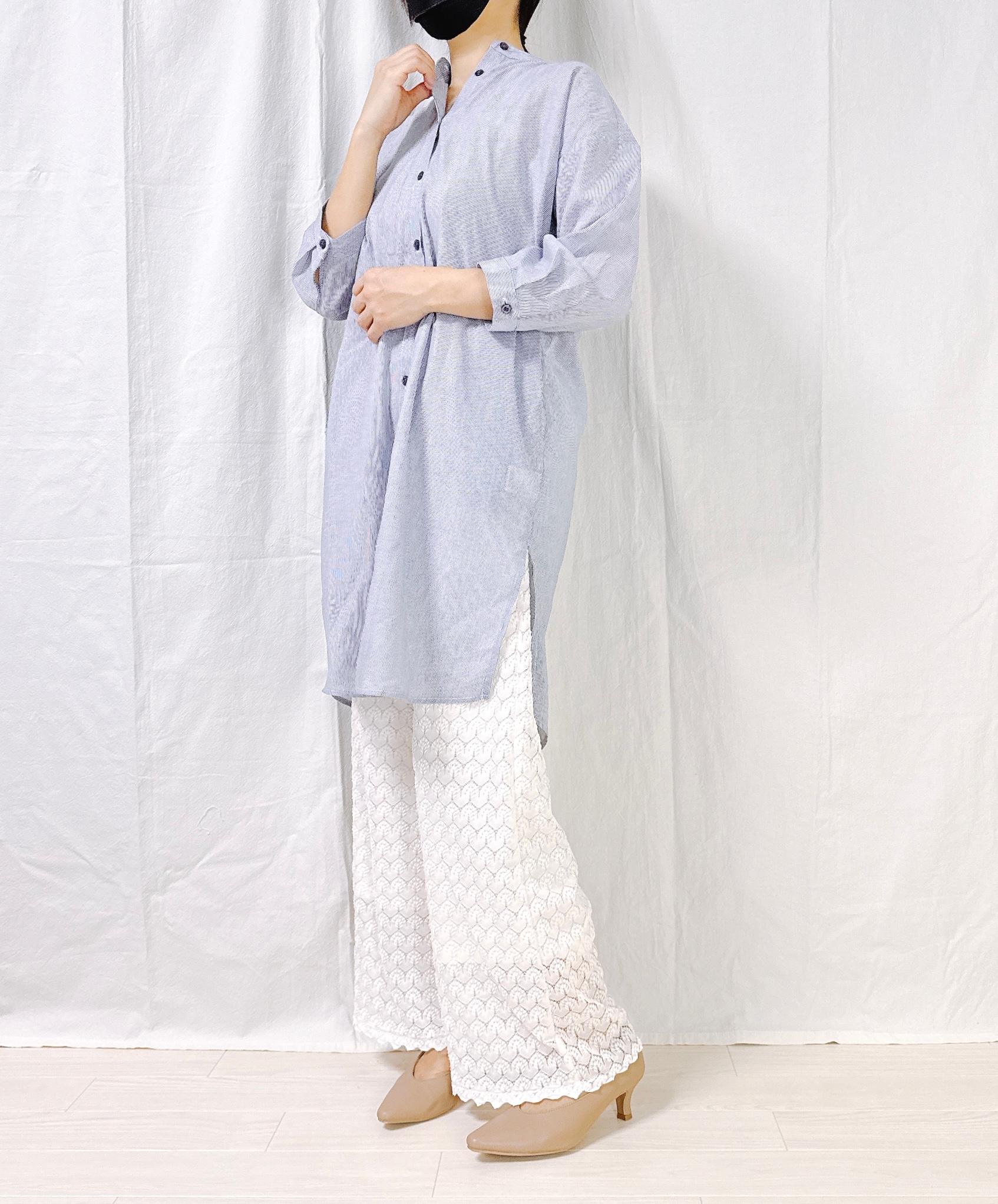 Person wearing a long, buttoned tunic over lace-patterned pants and heeled shoes, standing against a plain backdrop