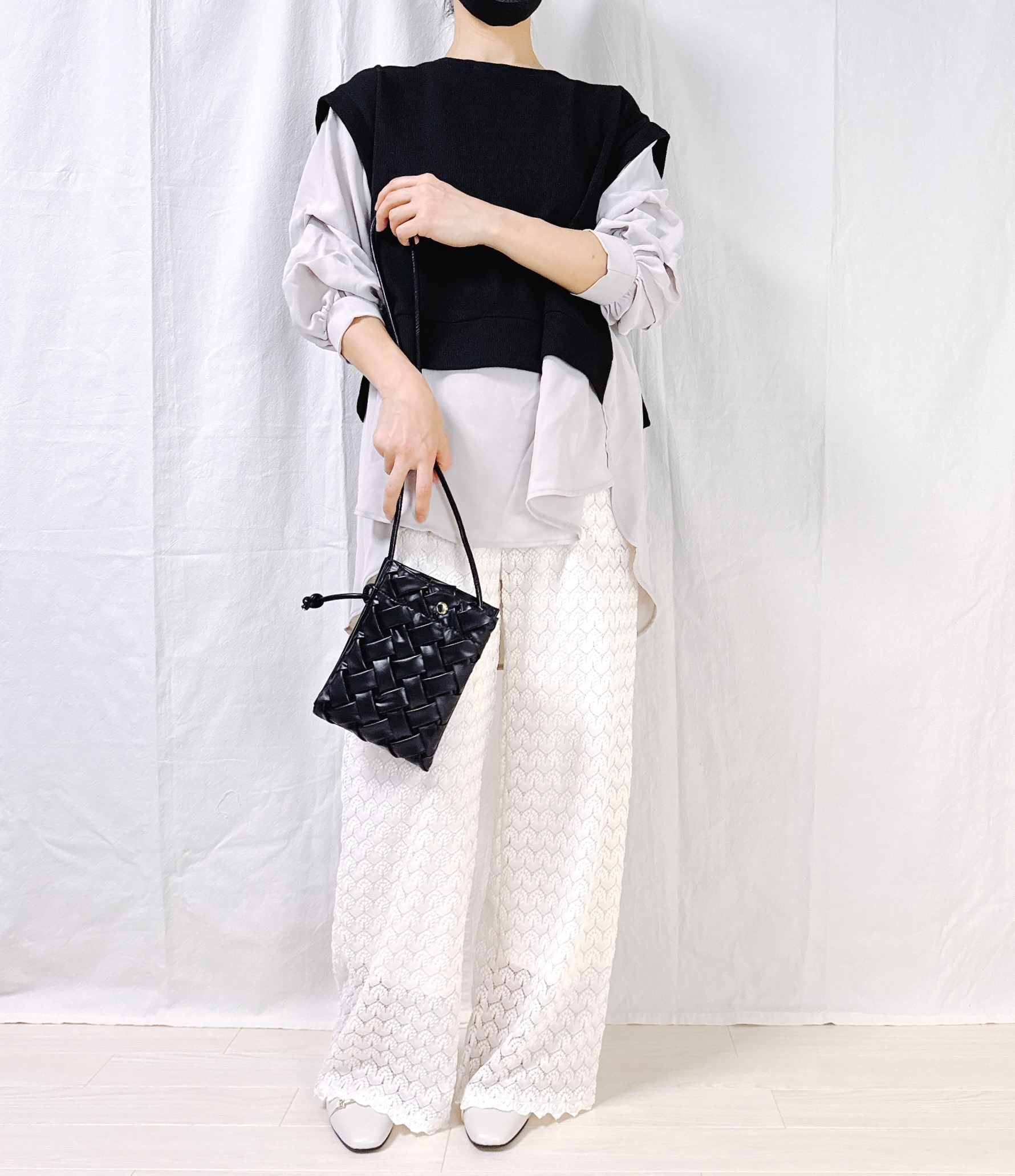 Person wearing layered clothing with a sleeveless black top, long-sleeve shirt, and white patterned pants, holding a woven handbag. Face is not visible
