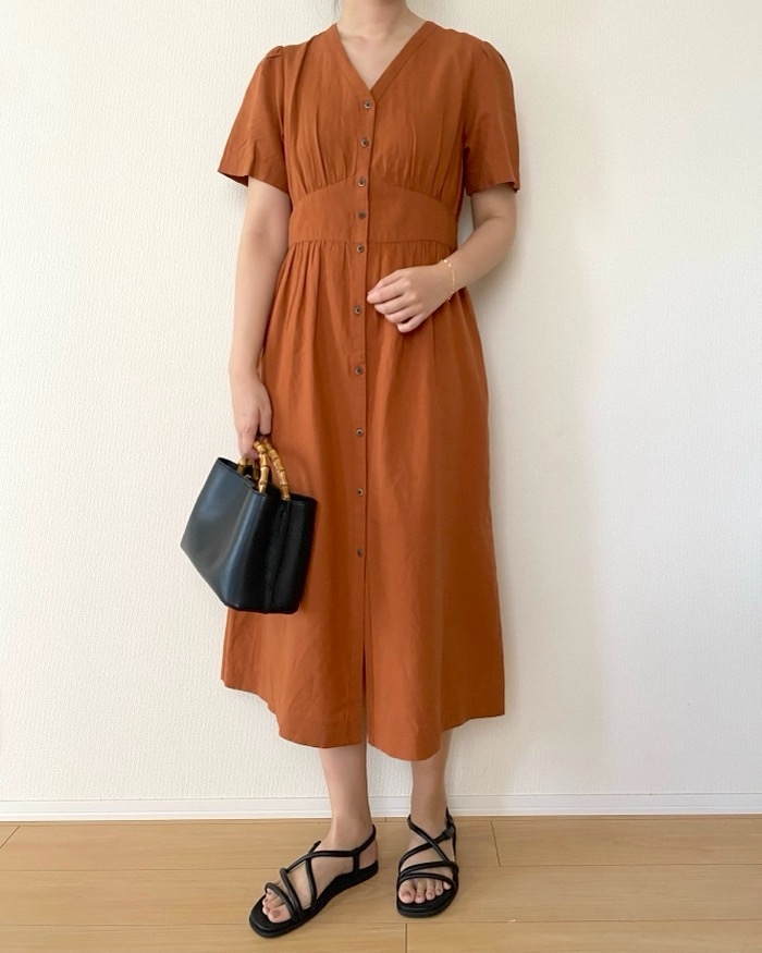 Person wearing a button-up dress holding a black bag with wooden handles. They are in sandals, standing against a plain background
