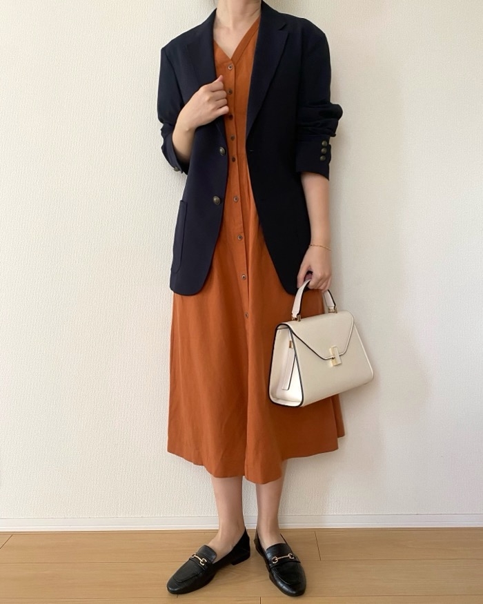 Person wearing a blazer over a button-down dress, holding a handbag, and wearing loafers, standing against a plain background