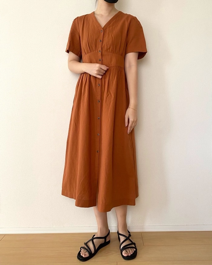 A person wearing a button-down, short-sleeved, knee-length dress and strappy sandals stands against a plain background