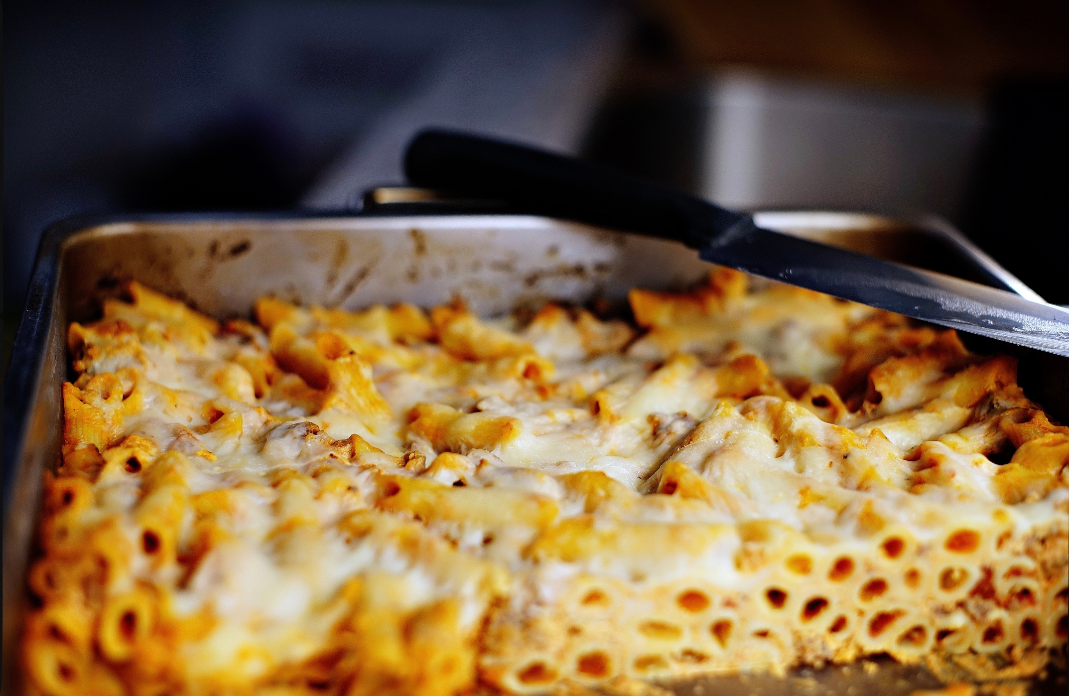 Pasta bake in a metal tray, partially sliced, with a serving spatula resting on the edge