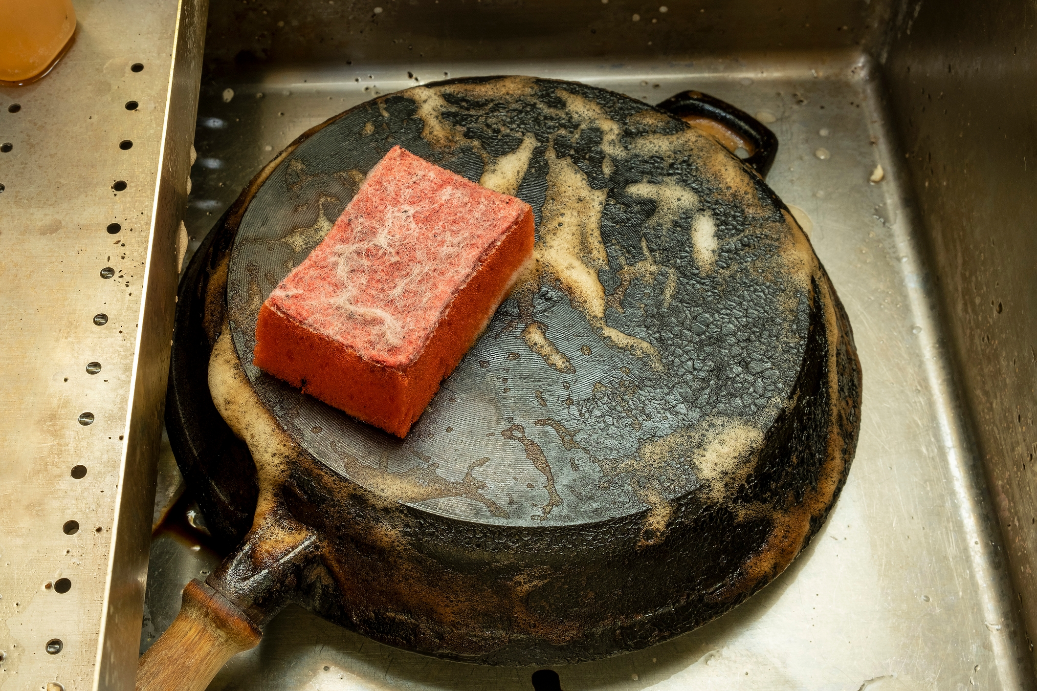 Dirty cast iron pan in a sink with soap suds and a red scrubber sponge on top