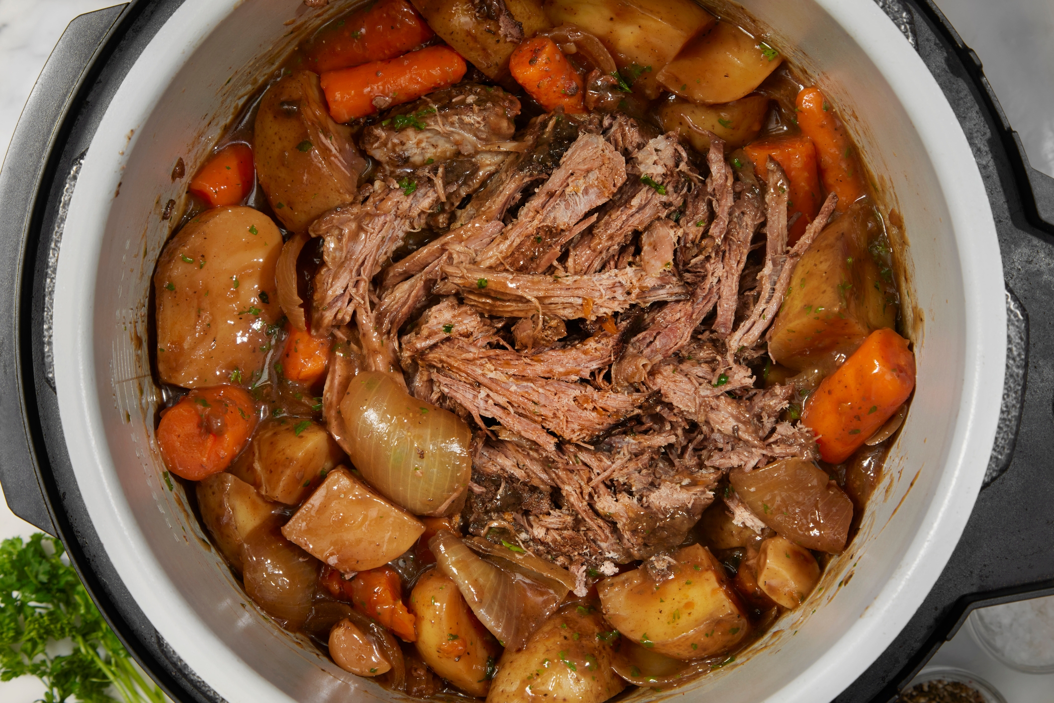Beef pot roast with carrots, potatoes, and onions in a slow cooker, garnished with herbs