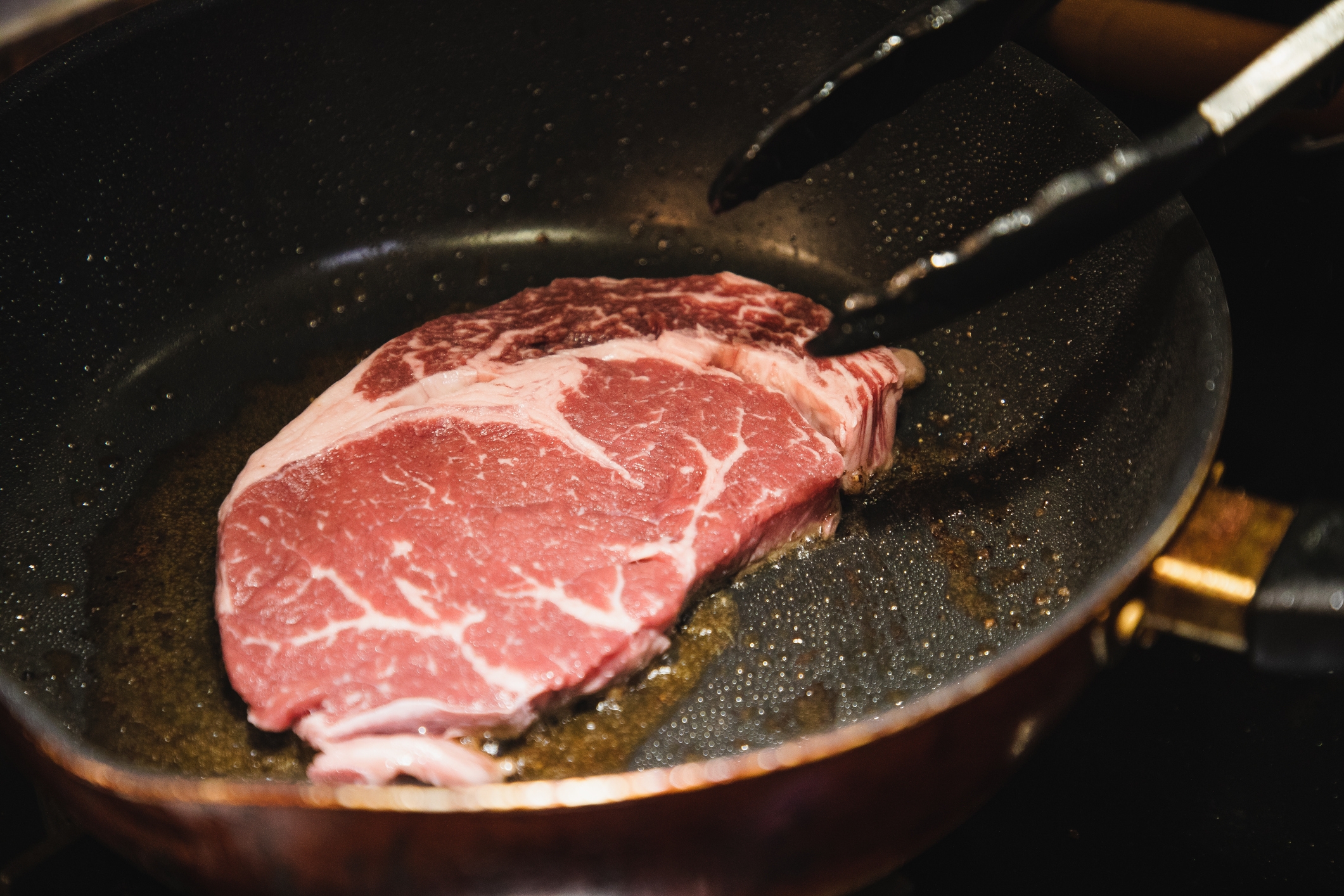 Steak cooking in a frying pan with tongs holding it. Visible marbling on the meat, indicating a cut with fat