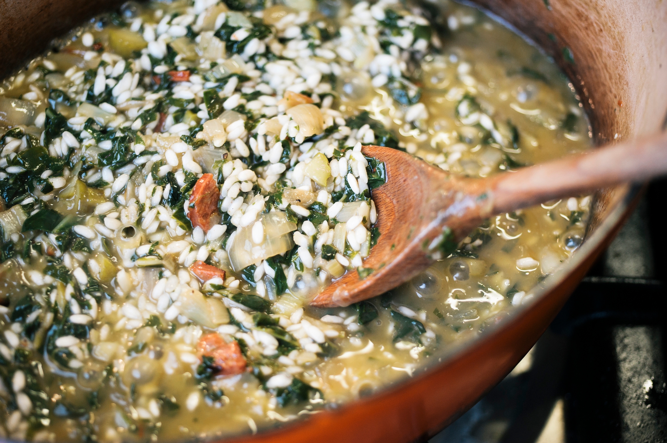 Close-up of a pot with risotto, including leafy greens and vegetables, being stirred with a wooden spoon