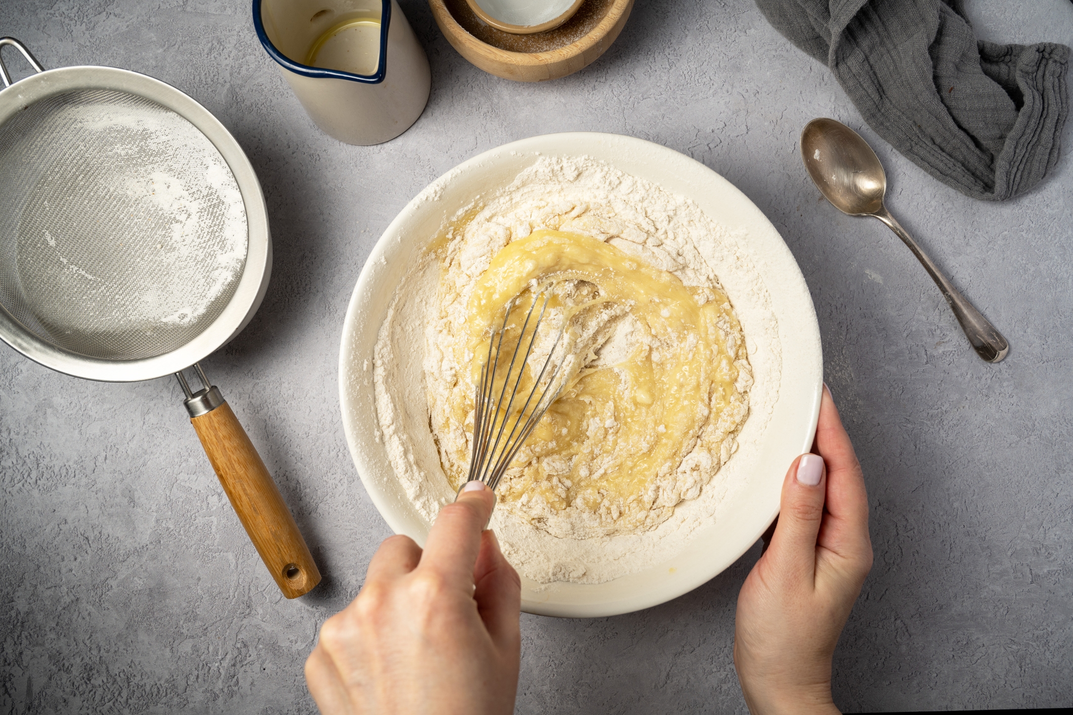 A person whisks batter in a bowl on a kitchen counter, surrounded by a sifter, spoon, and cloth