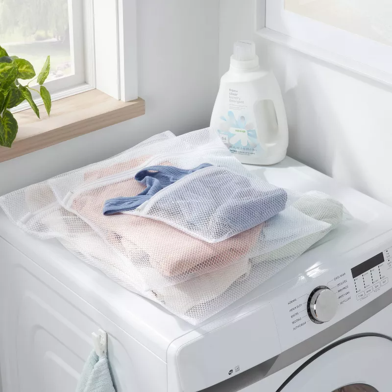 Laundry items in mesh bags sit on a washing machine next to detergent, near a plant on a window ledge