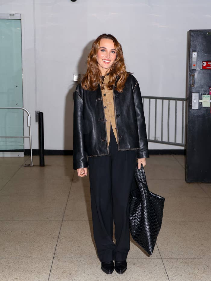 Person in a buttoned shirt and leather jacket, wide-leg pants, holding a large woven tote bag, standing indoors