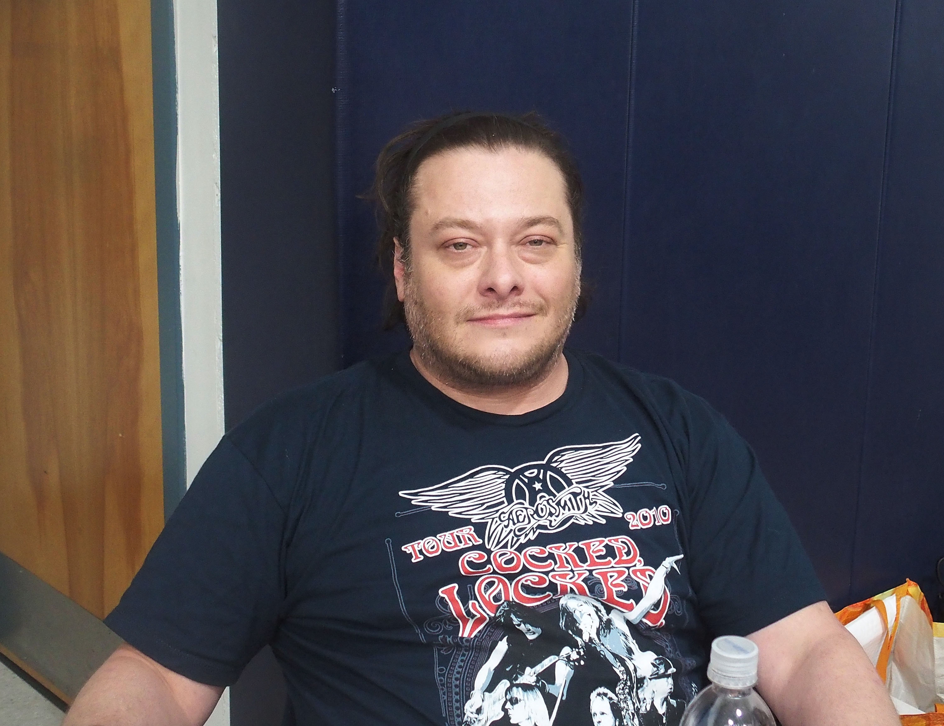 A person sits indoors, smiling, wearing a black graphic T-shirt with an Aerosmith logo and the words "2010 Cocked Locked."