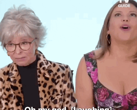 Two women sit side by side; one in a leopard print jacket and glasses, the other in a floral dress, both reacting with surprise and laughter