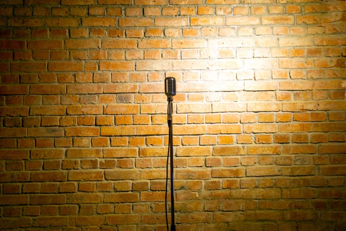 A vintage microphone stands alone in front of a brick wall, suggesting a setting for a live performance or stand-up comedy show