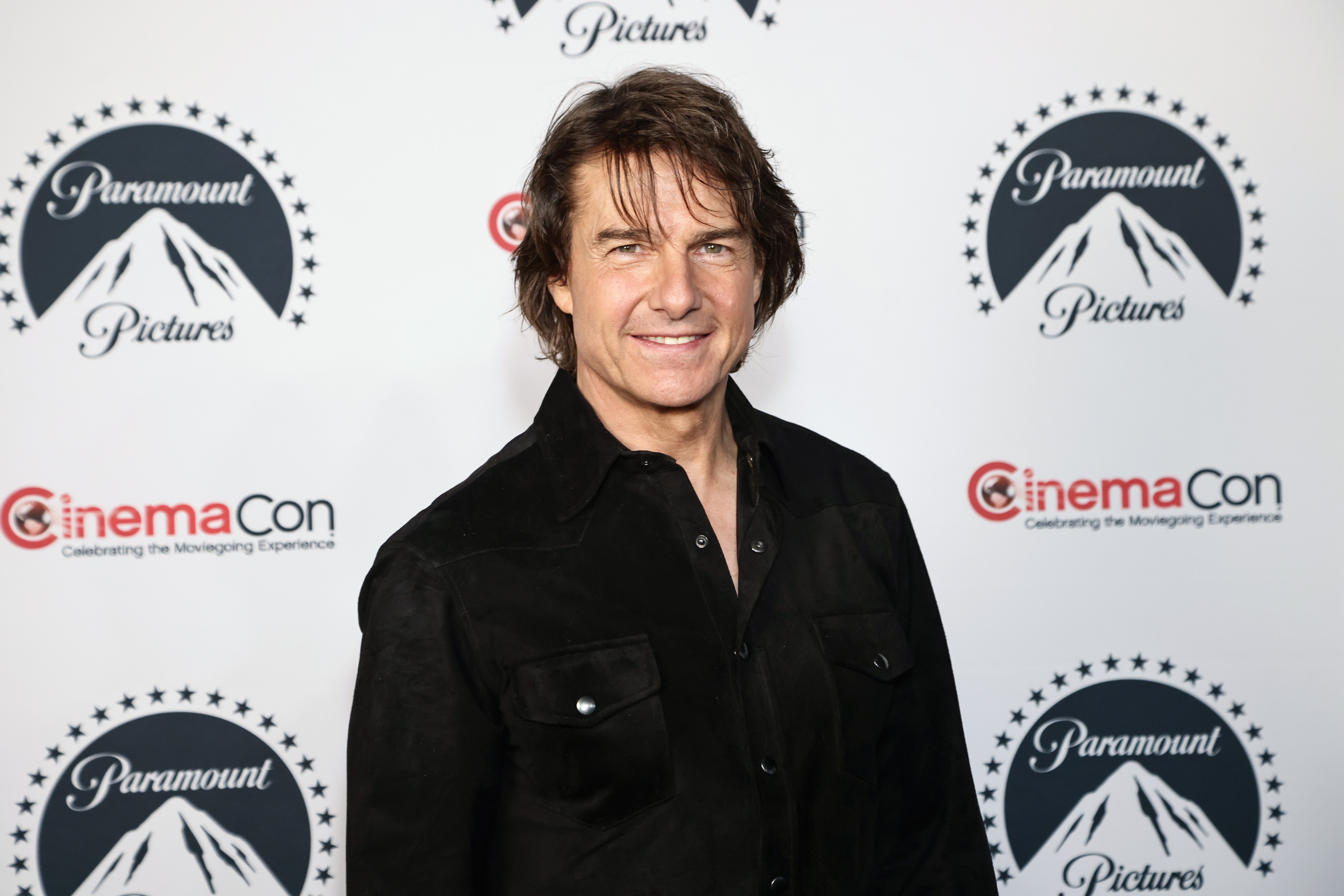 Tom Cruise at CinemaCon event, wearing a button-up shirt, stands in front of a Paramount Pictures backdrop, smiling at the camera