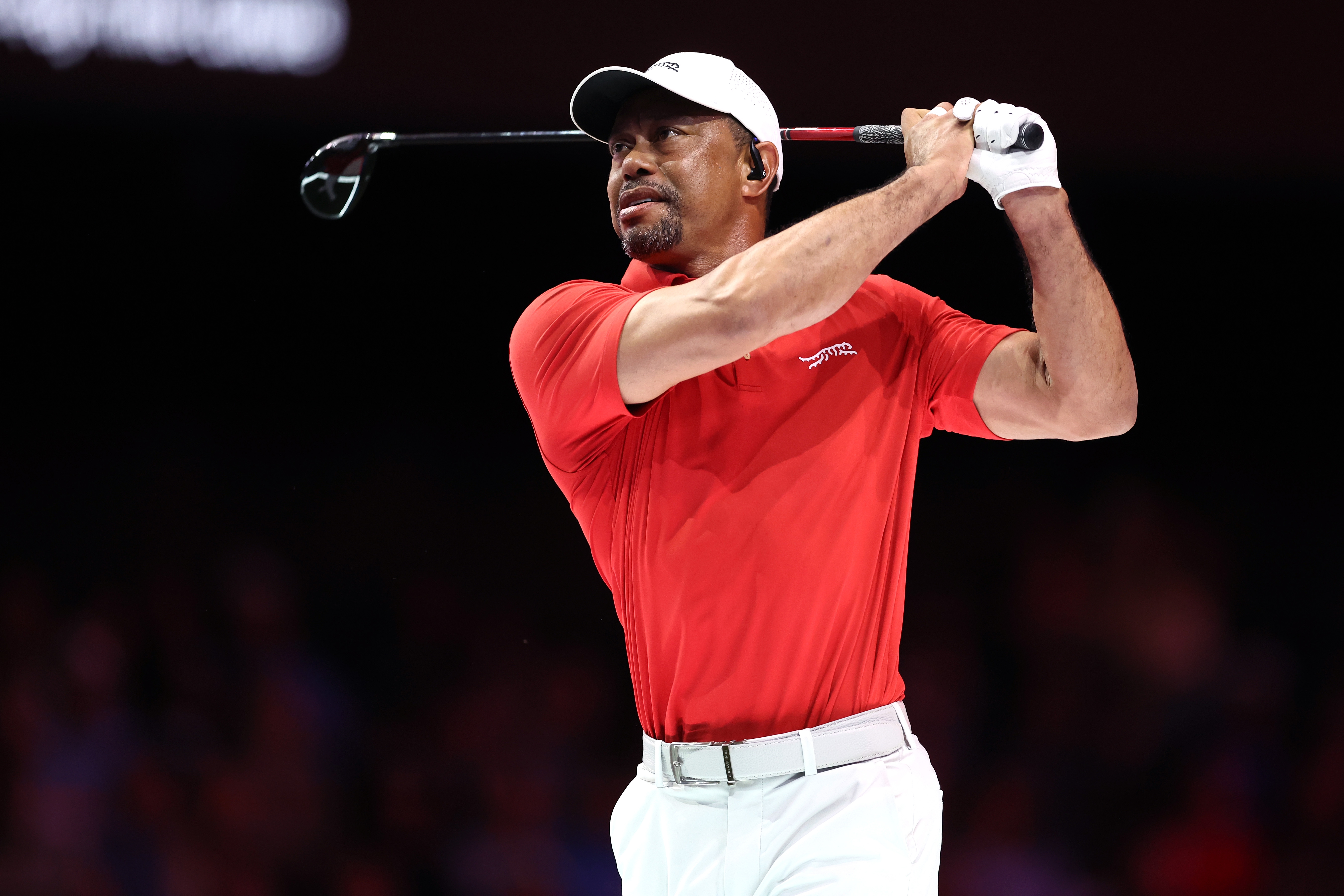Tiger Woods in a red polo and white cap finishes a swing, focused and poised, on a dimly lit course