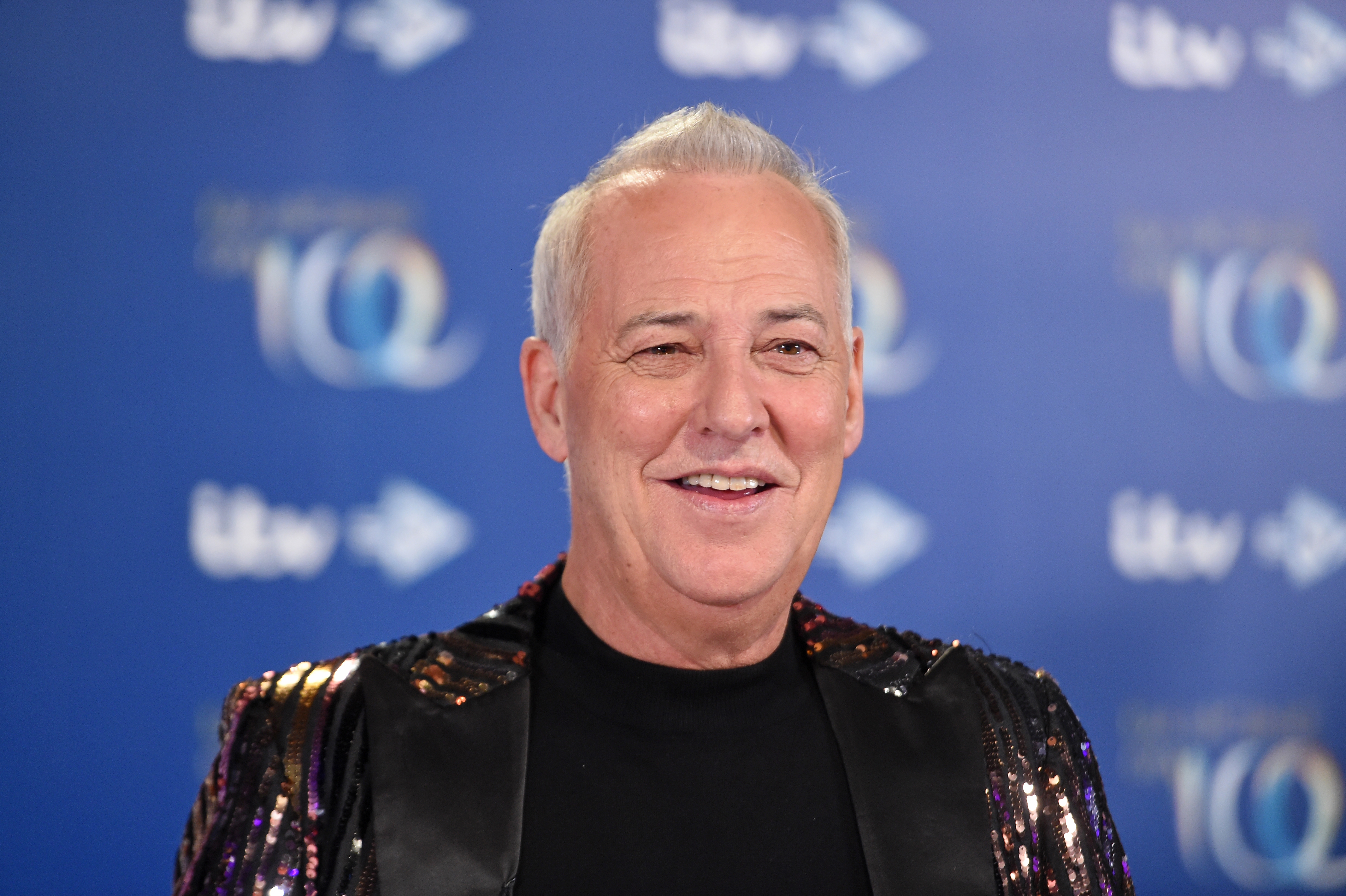 Michael Barrymore smiles on a blue TV event backdrop, wearing a shiny, sequin-covered jacket over a black top