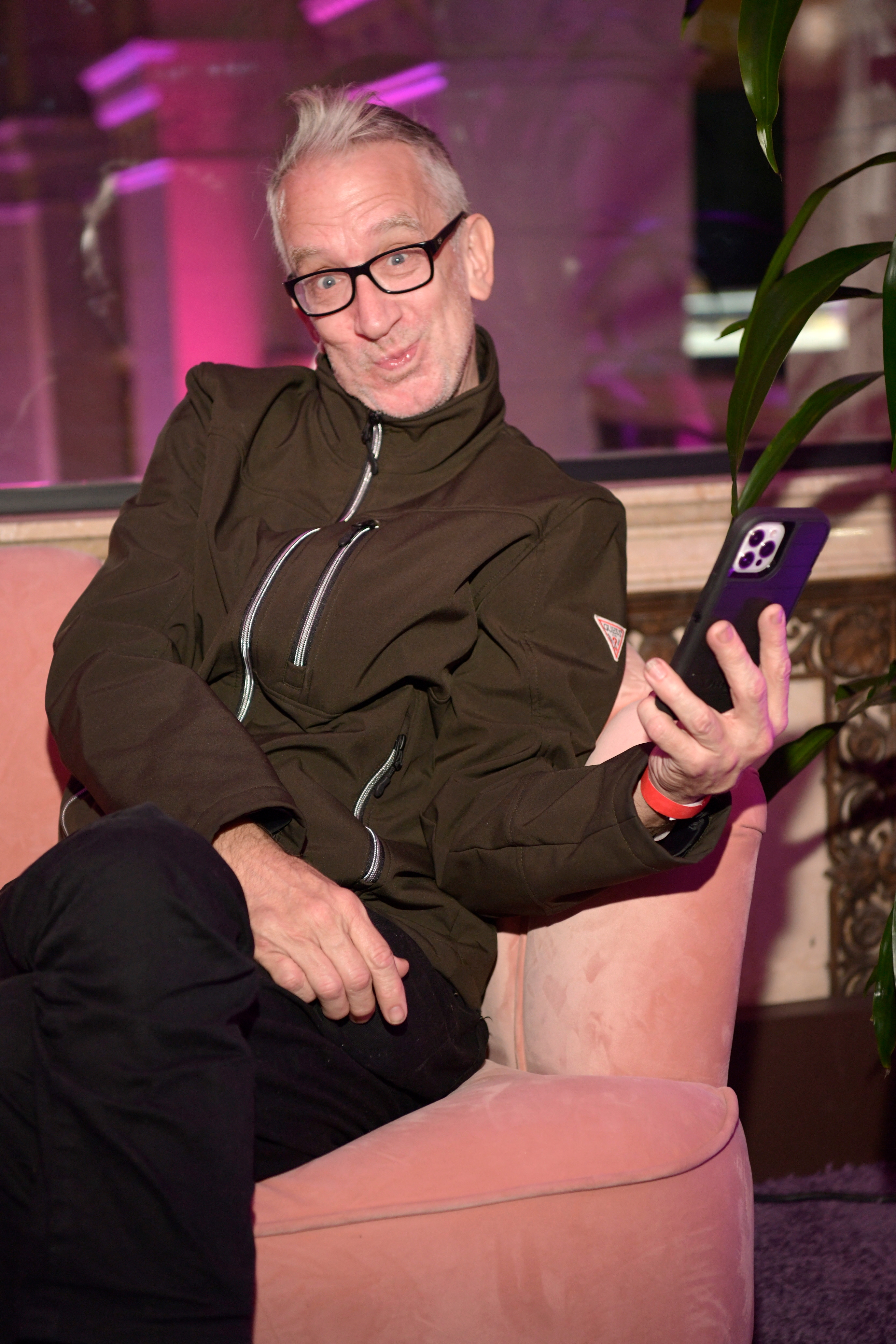 Andy Dick in a casual jacket sits on a plush chair, smiling and holding a smartphone, with one leg crossed