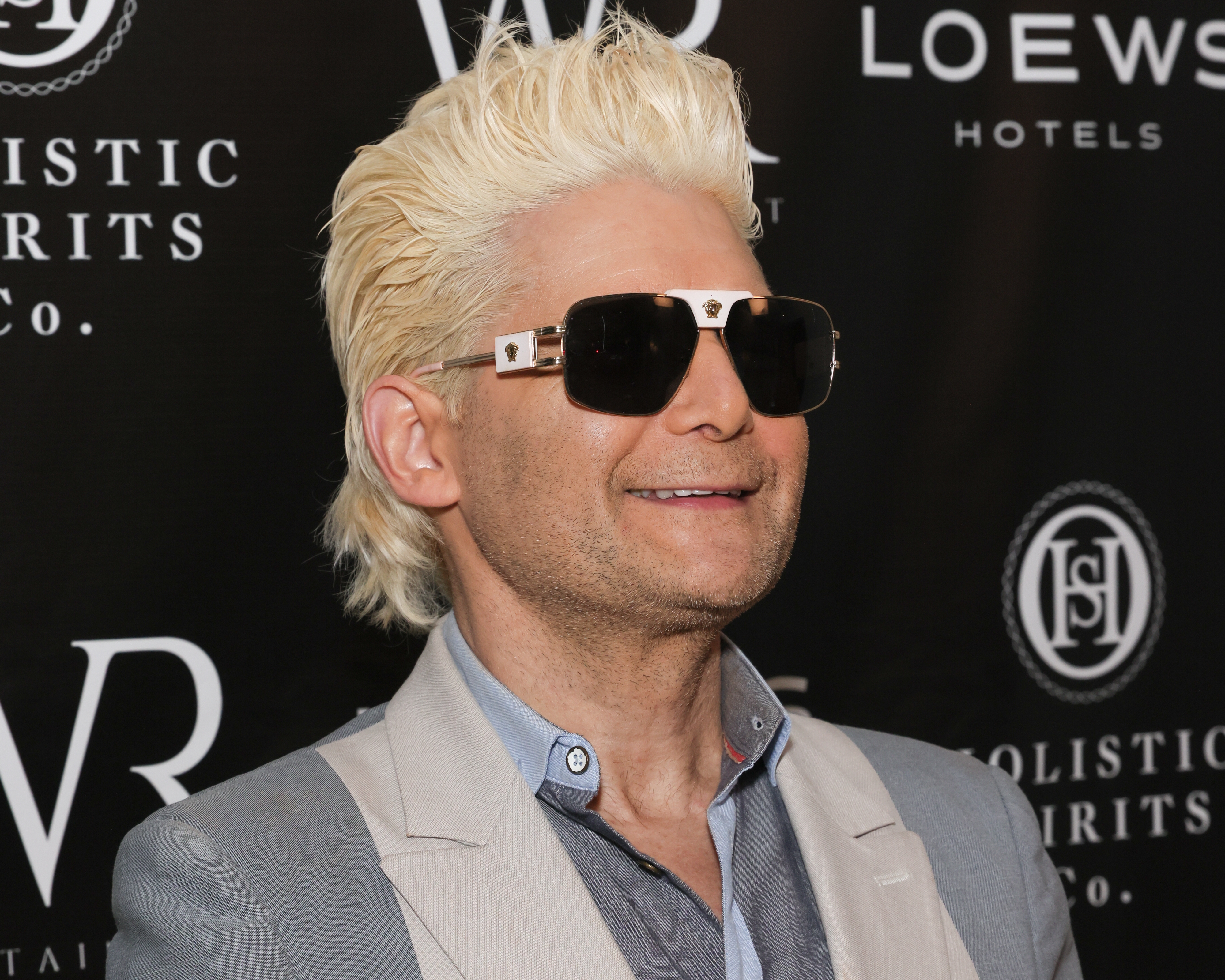 Corey Feldman with short, styled hair wearing sunglasses and a light-colored suit jacket on a media backdrop