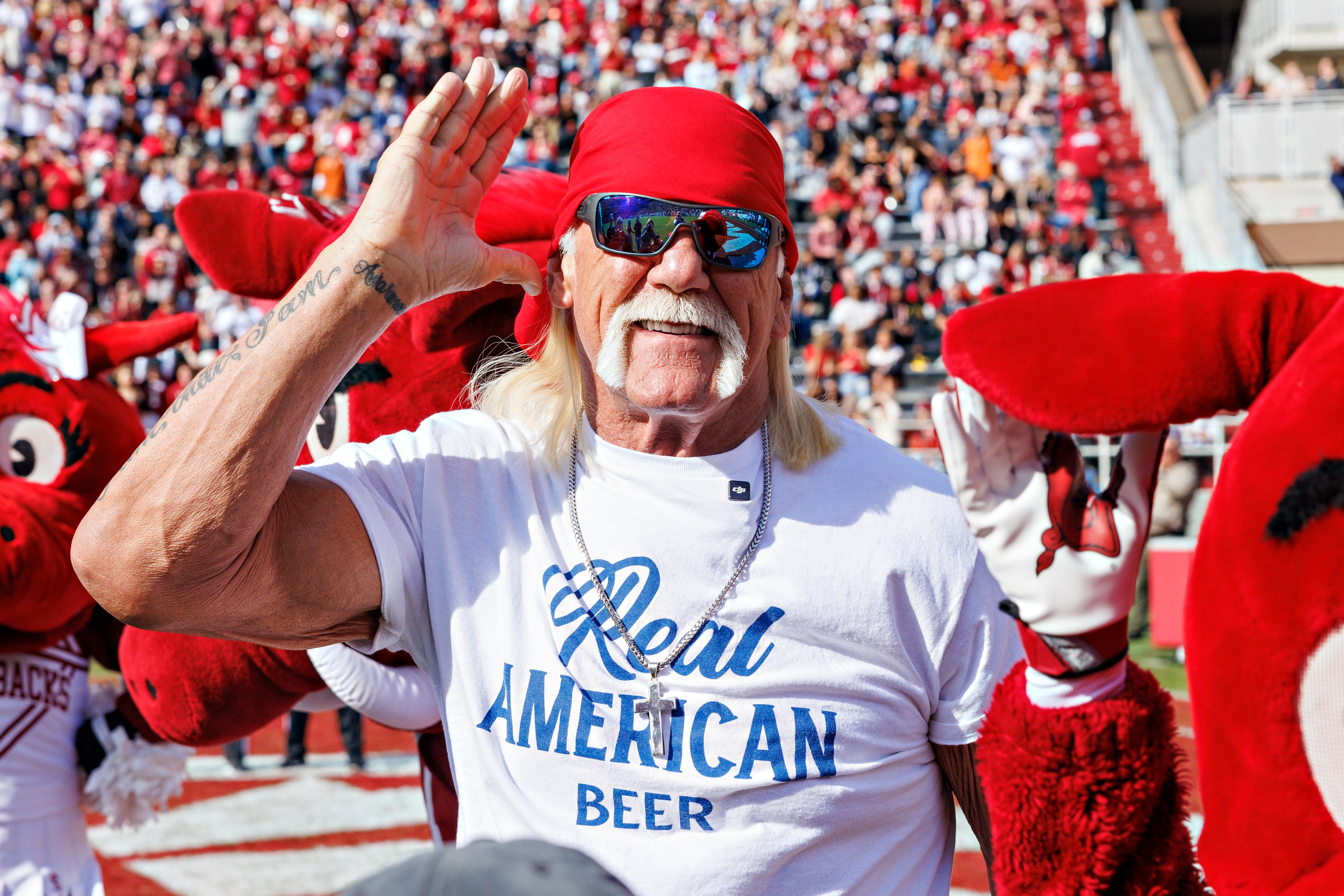 Person in a bandana and sunglasses with mascots, cupping hand to ear at outdoor event, wearing "Real American Beer" t-shirt