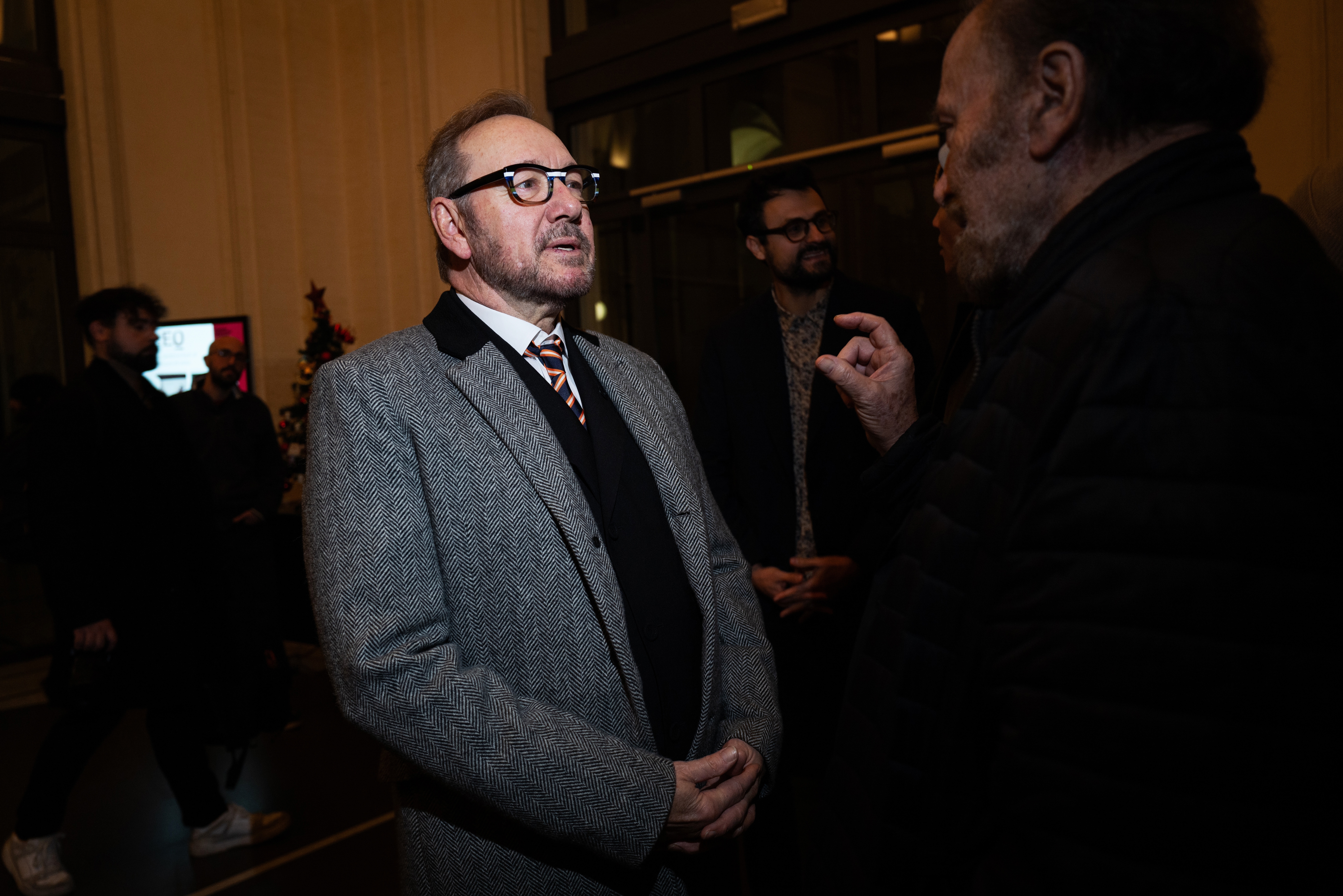 I don't know who this is, but the man wears a gray tweed coat and glasses while talking to another person in a dimly lit room