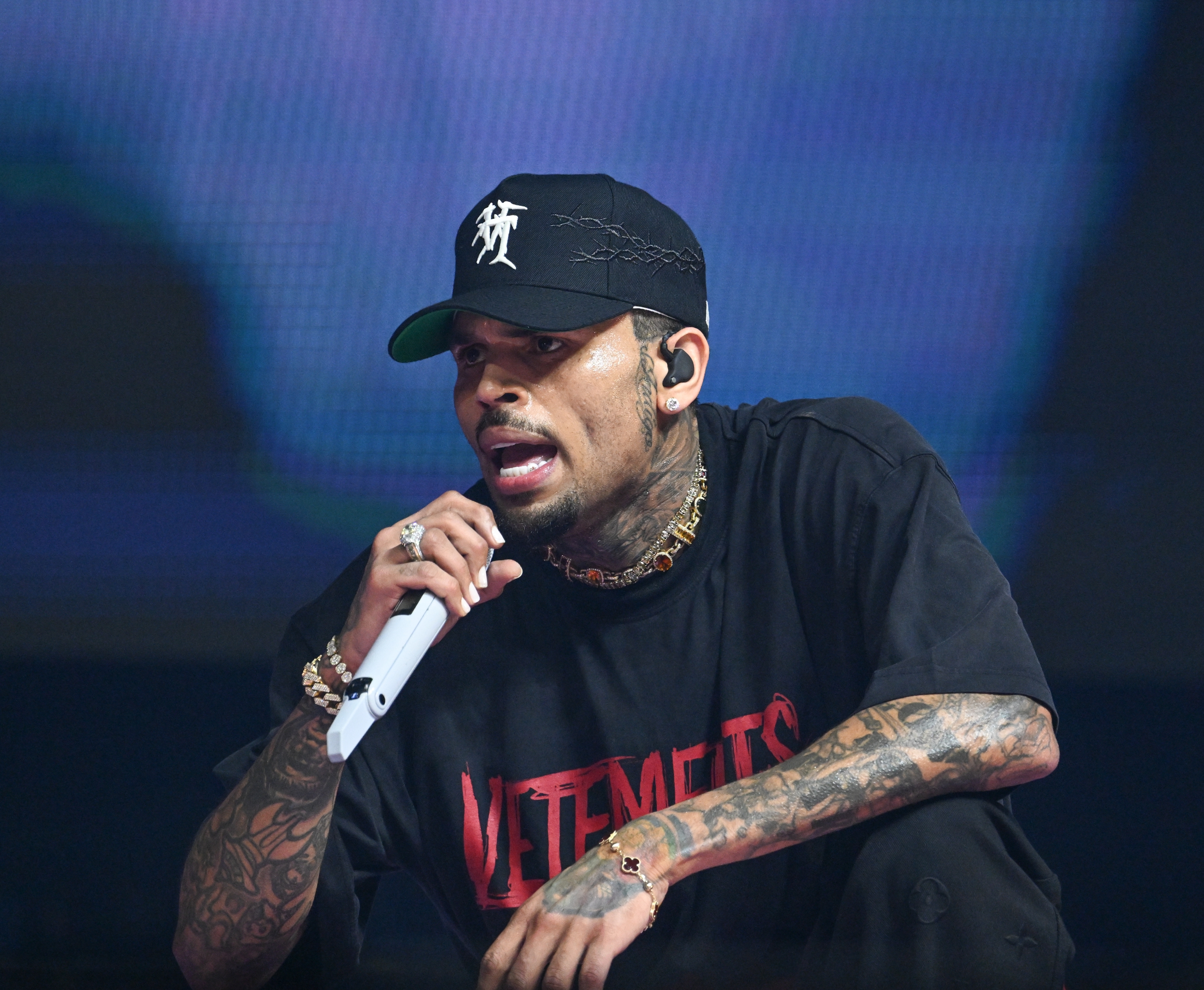 Chris brown on stage wearing a black cap, graphic t-shirt, and jewelry, holding a microphone and performing passionately