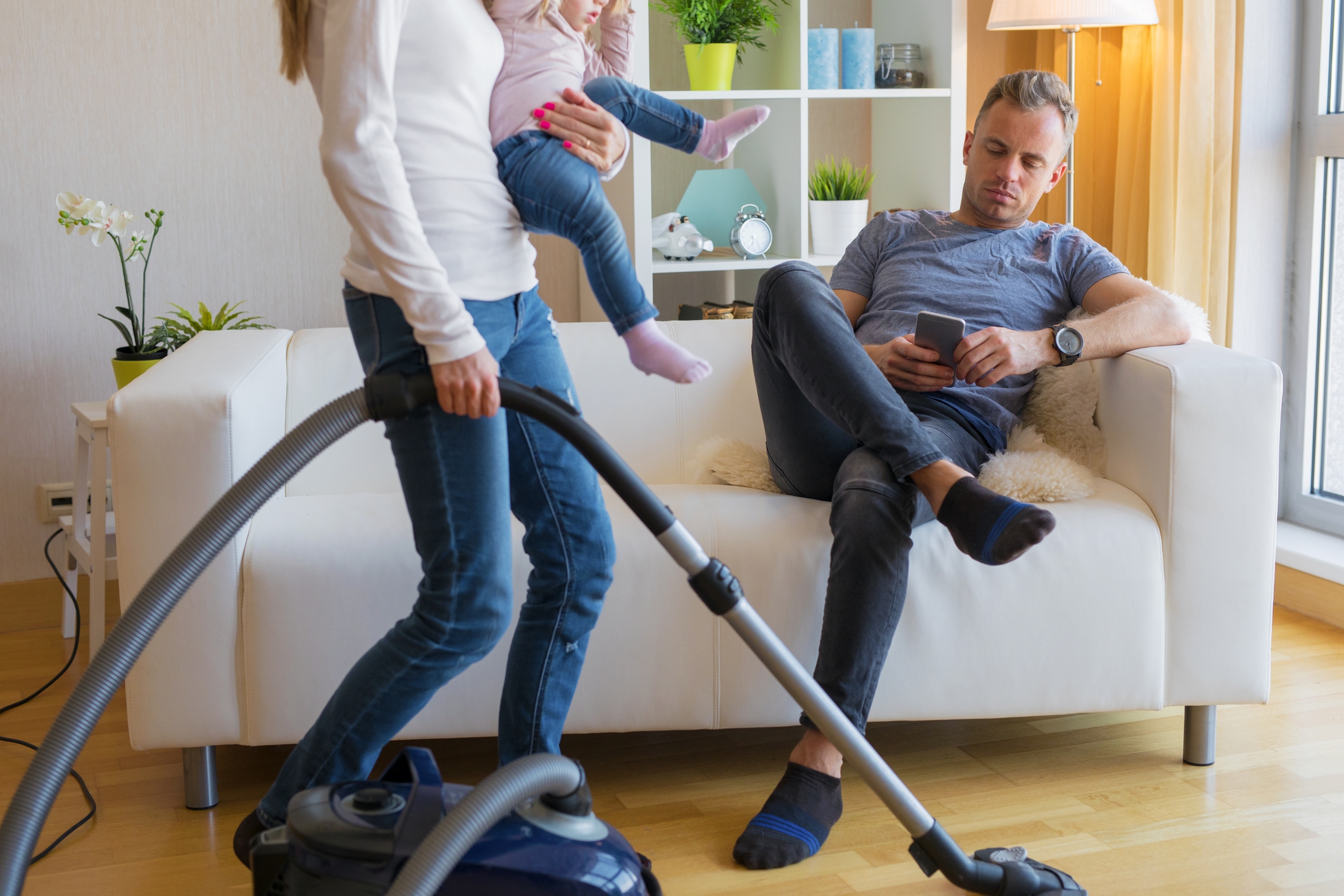 Man sitting on a couch using a phone, while a woman vacuums holding a child, in a living room setting