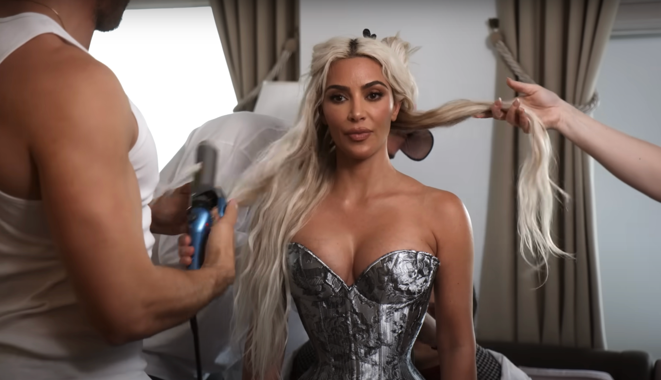Person with long hair styled by a team, wearing a metallic corset dress in what appears to be a preparation setting