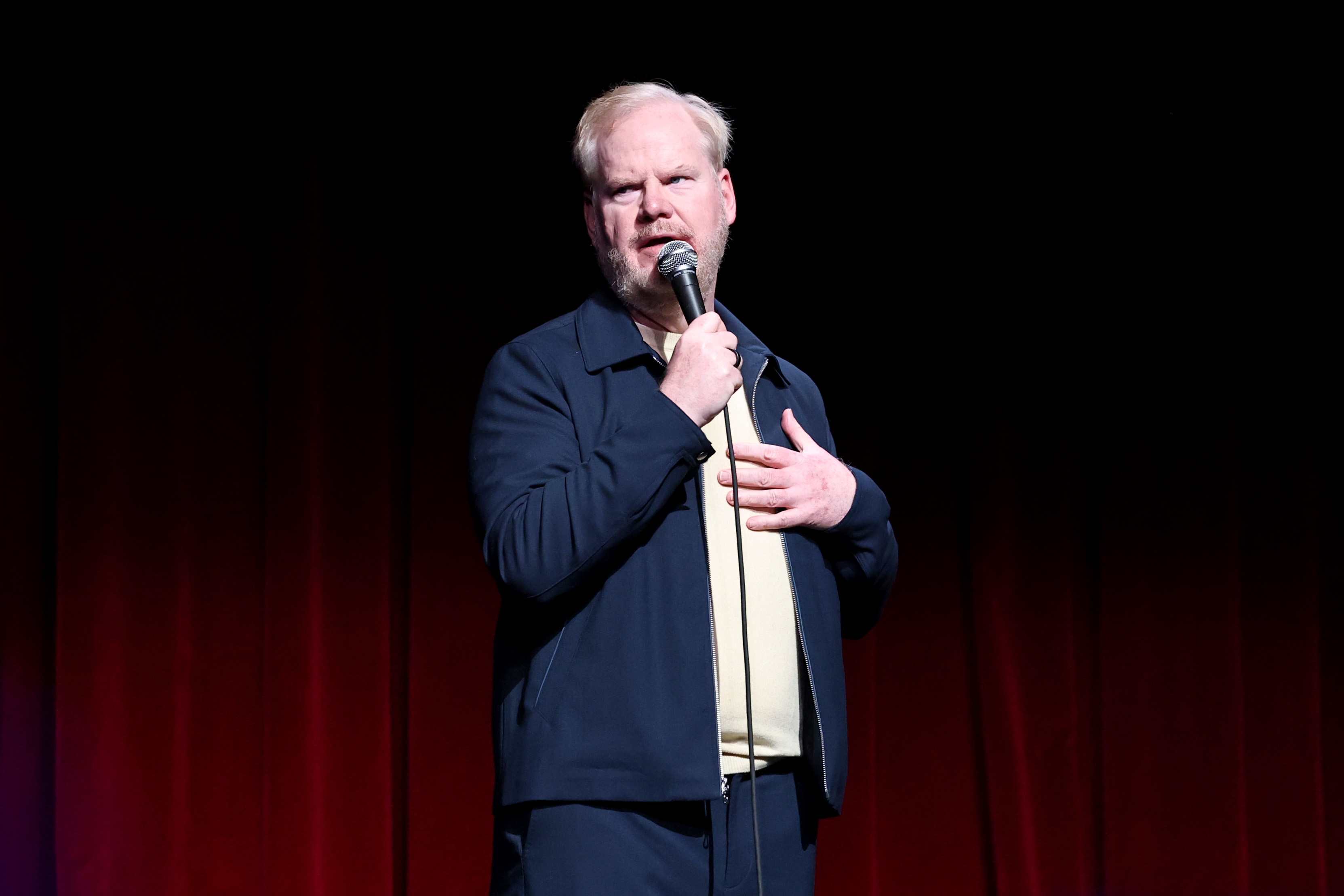 Jim Gaffigan performing on stage, holding a microphone, wearing a casual jacket over a light-colored shirt. Dark background with red curtains