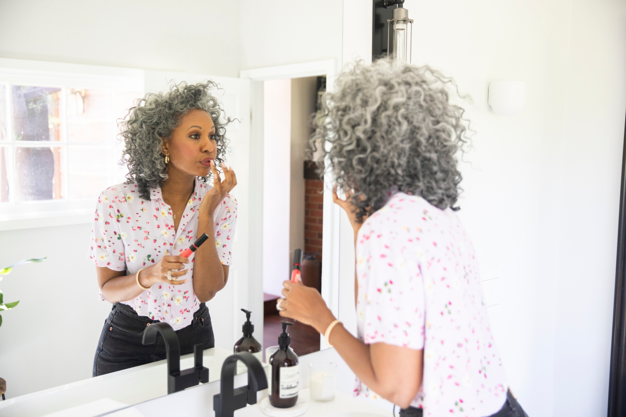 A person with curly hair applies lipstick in front of a bathroom mirror, wearing a floral-patterned shirt and dark pants