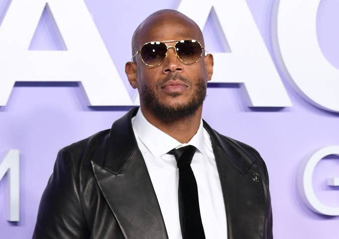 Person on the red carpet in a leather jacket, white shirt, black tie, and sunglasses, posing with hands in pockets