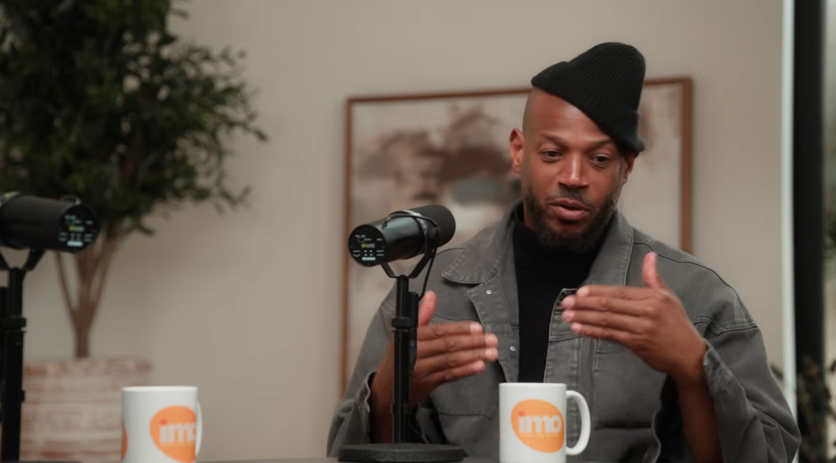 Person in a podcast studio wearing a casual jacket and beanie, gesturing while speaking into a microphone