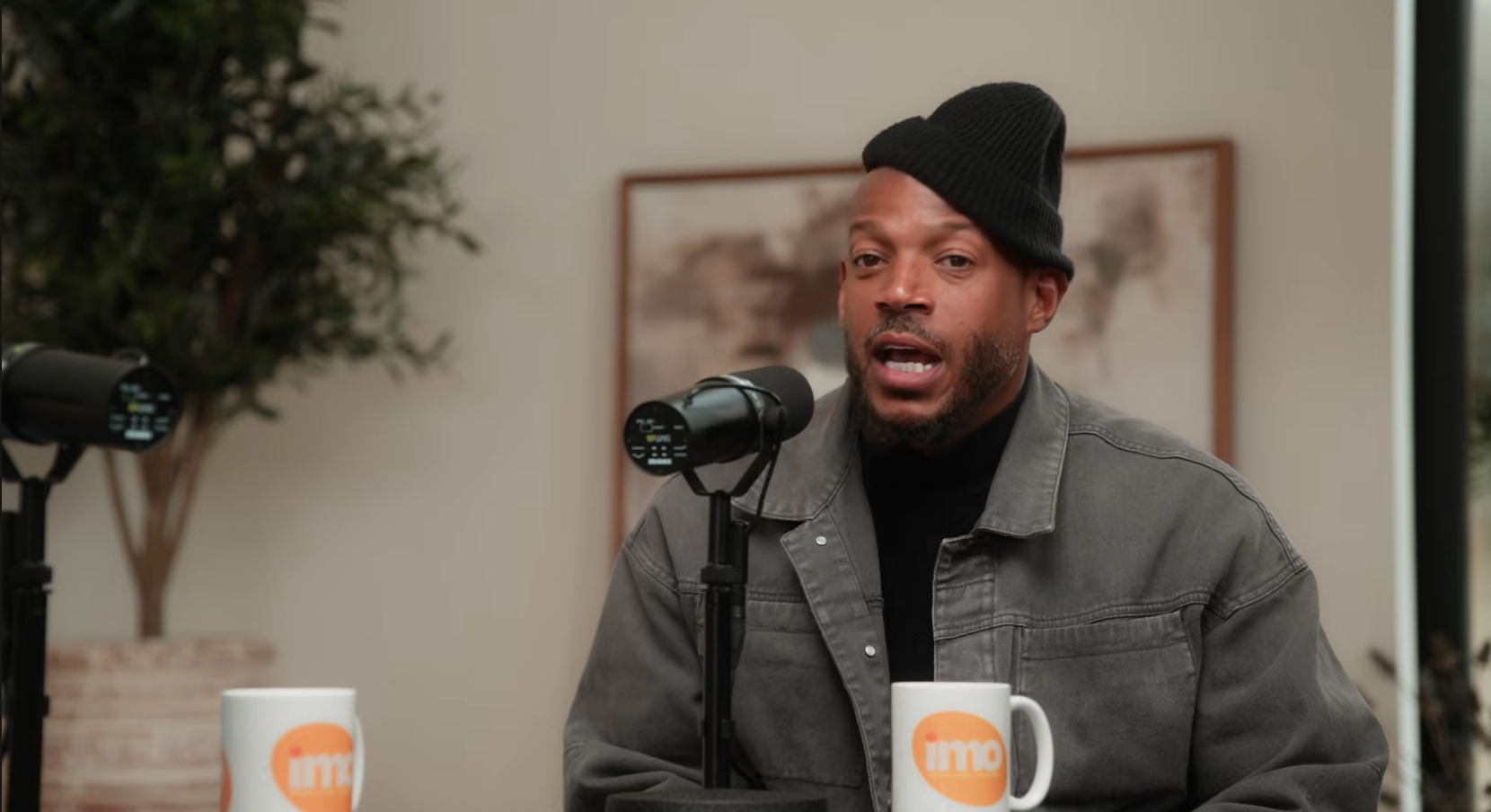 Person in a casual jacket and beanie speaking into a microphone during a podcast interview, with studio decor in the background