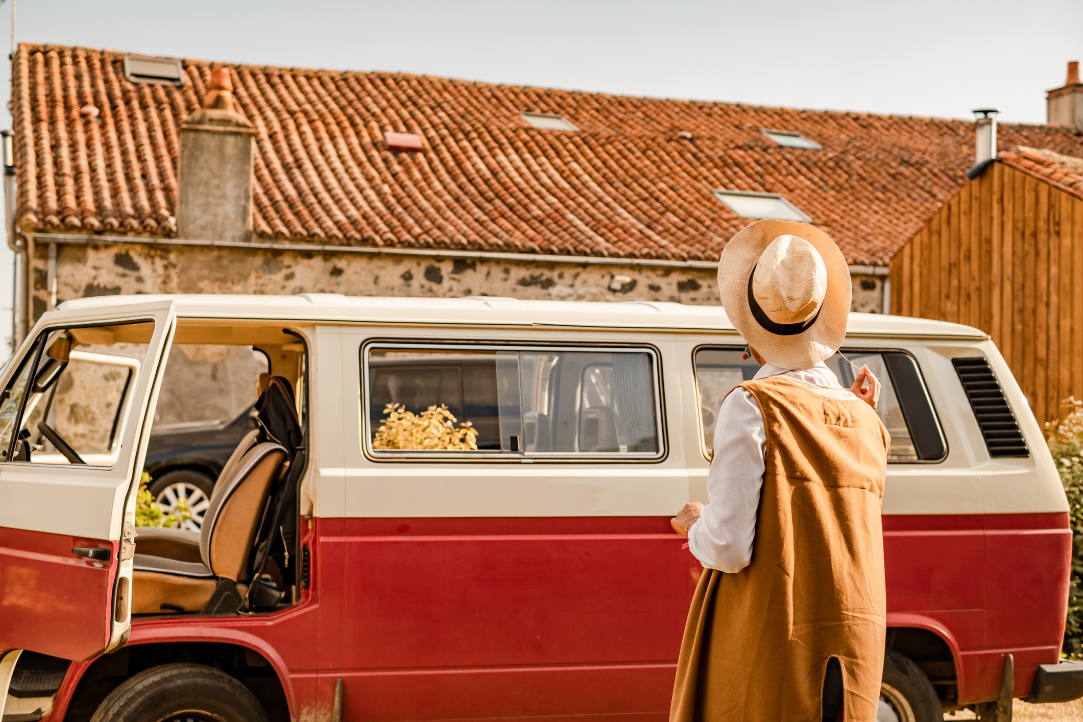 Person in vintage outfit with hat stands near classic van parked by rustic buildings