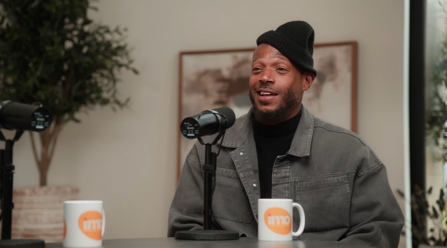 Person in a casual jacket and beanie sits talking into a microphone during a podcast recording
