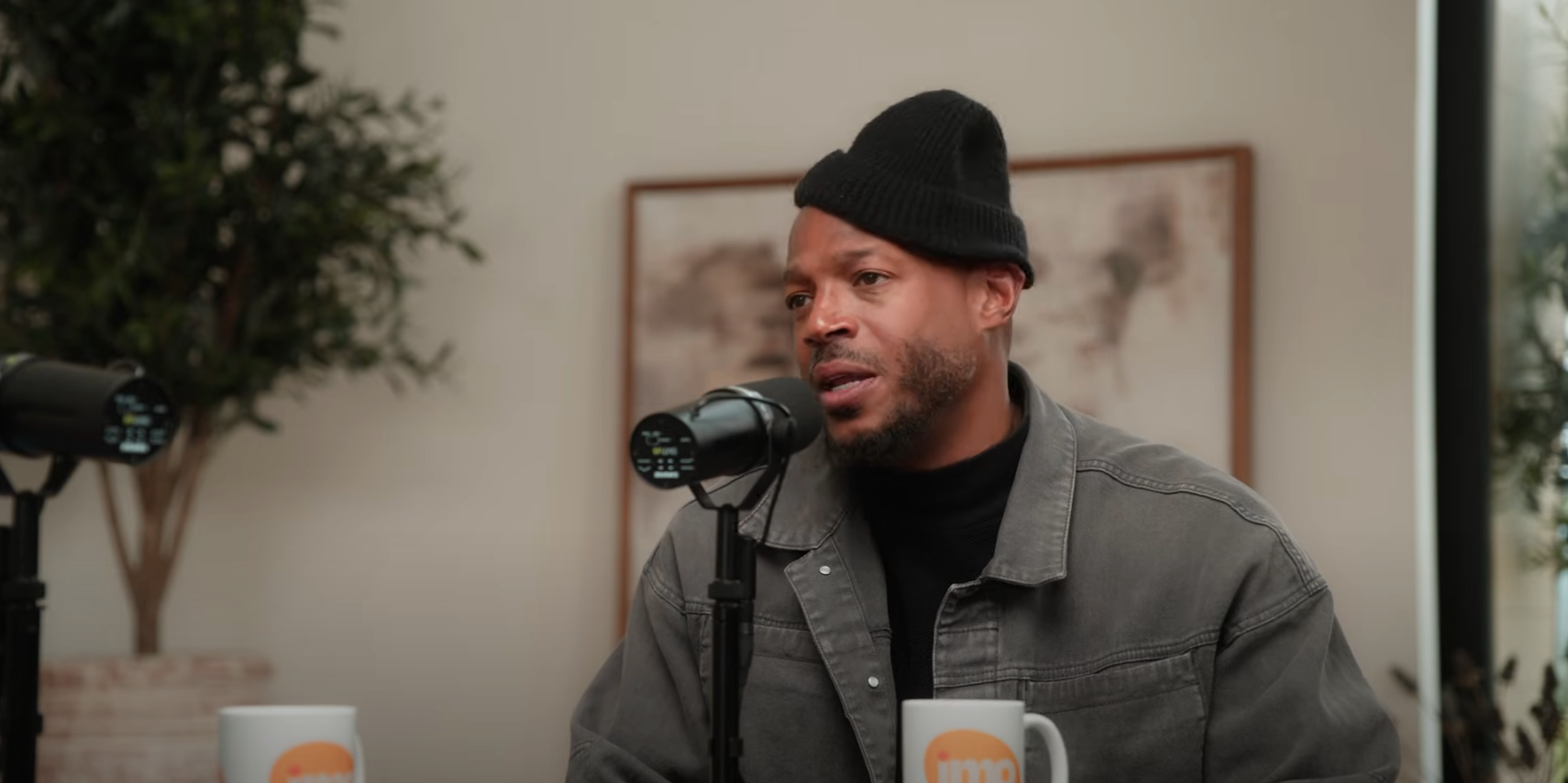 Person speaking into a microphone during a podcast, wearing a casual jacket and beanie. Coffee mugs and a blurred plant are in the background